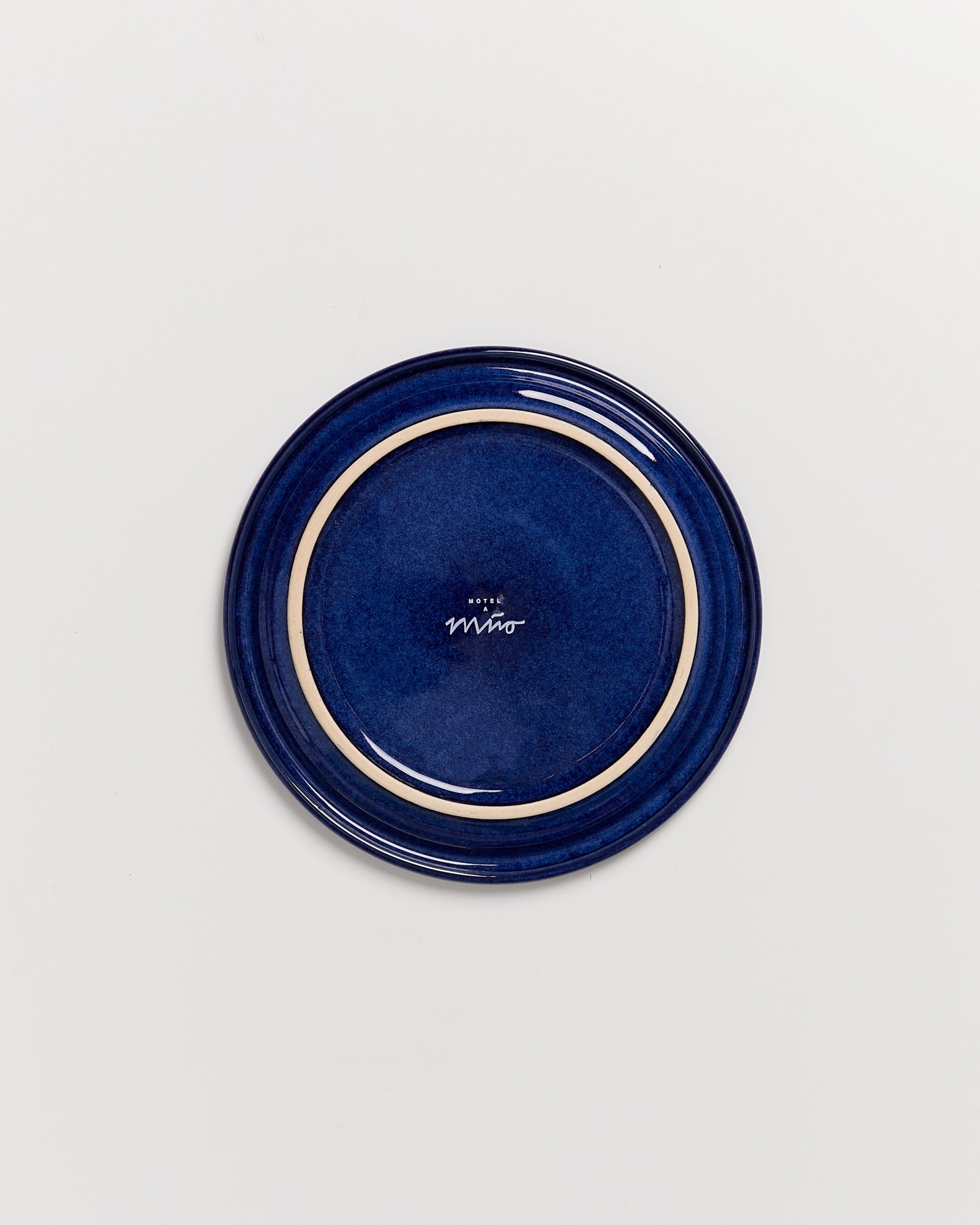 Rückseite eines blauen Tellers mit cremefarbenem Rand und einem Logo in der Mitte, auf weißem Hintergrund.