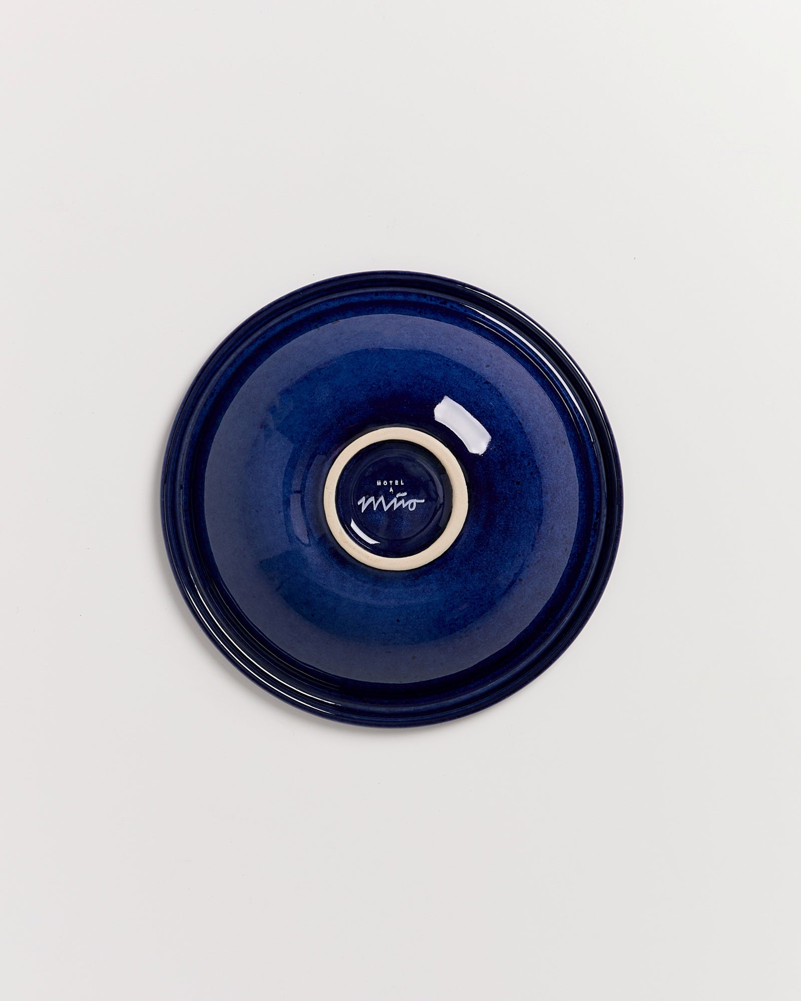 Blauer Keramikteller mit einem Signaturlogo in der Mitte, auf weißem Hintergrund.