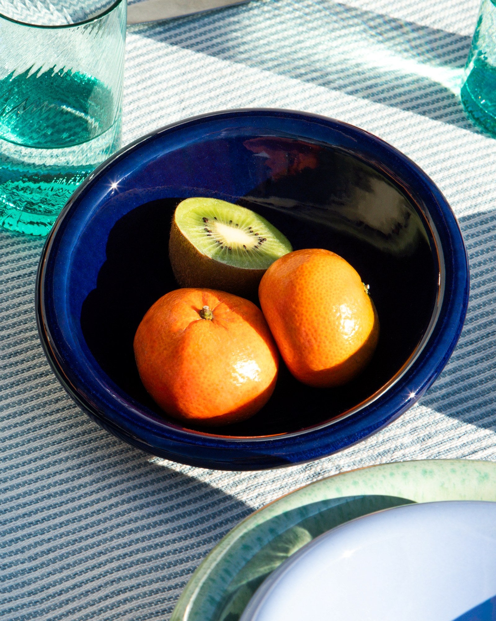 Eine blaue Schale mit zwei Mandarinen und einer halbierten Kiwi, daneben ein Glas Wasser auf einem gestreiften Tuch.