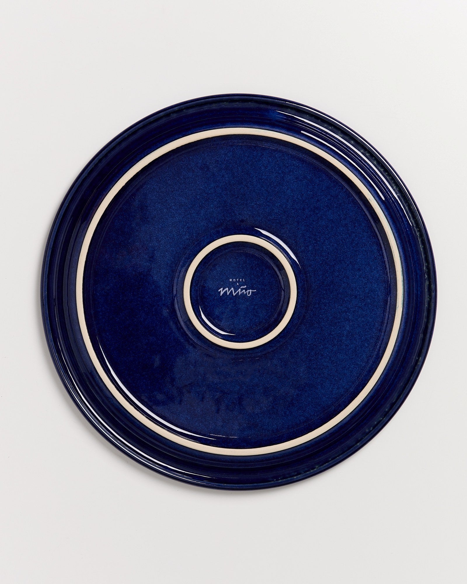 Dunkelblauer Teller mit beigem Rand und Aufschrift "Hotel Miro" auf der Unterseite, gegen weißen Hintergrund.