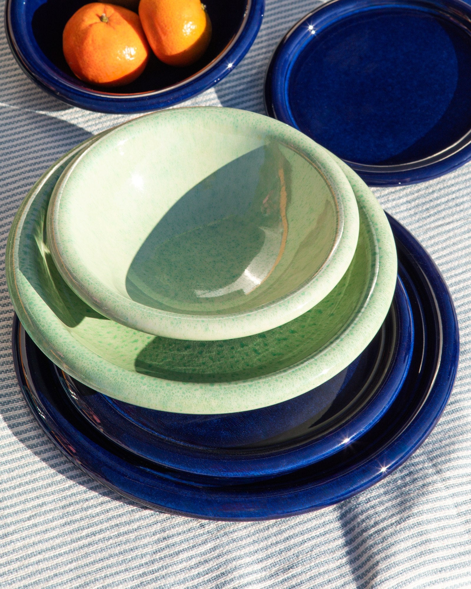 Ein Stapel aus grünen und blauen Keramiktellern und -schüsseln auf einem gestreiften Tuch, daneben Schale mit Orangen.