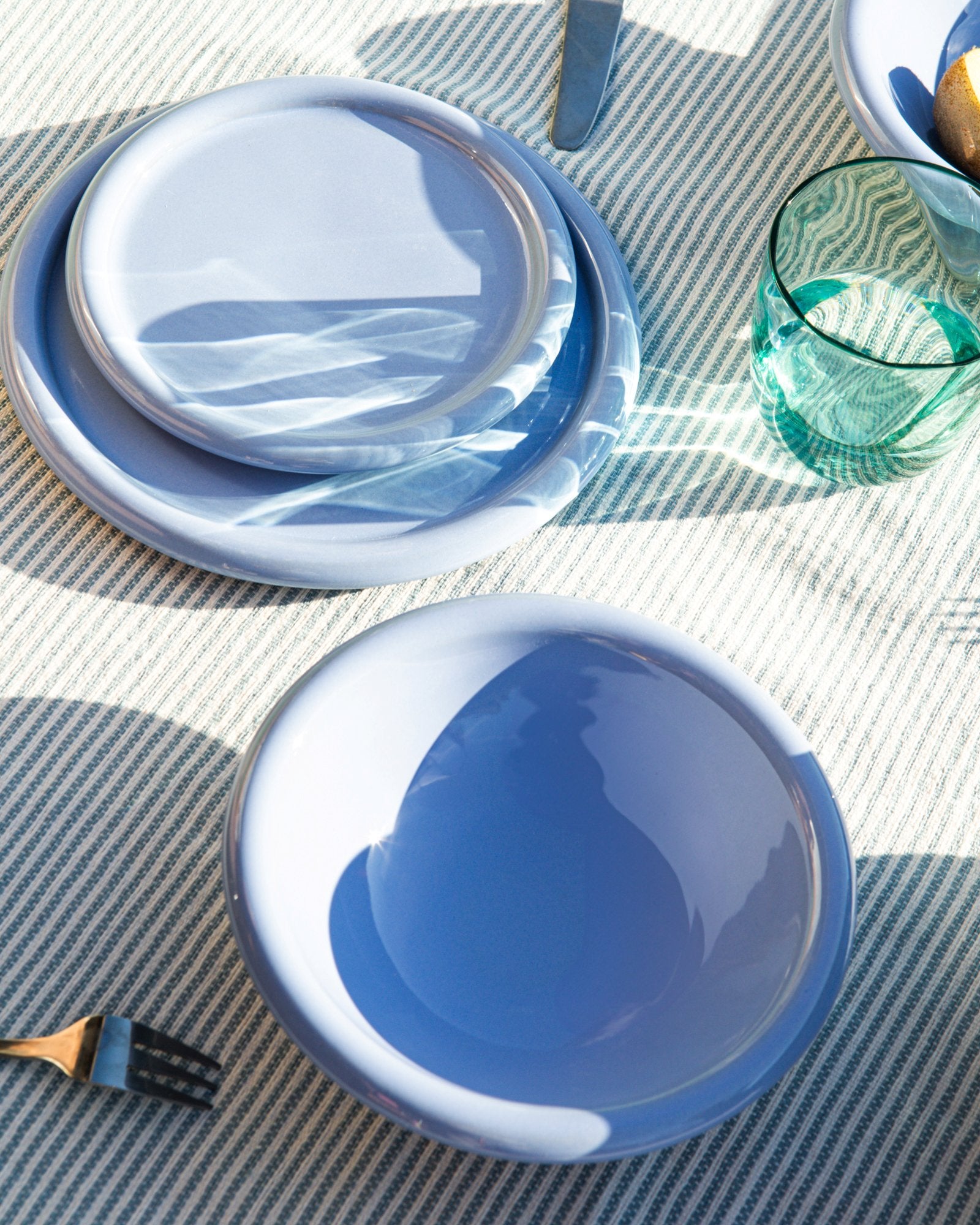 Blaue Teller und ein grünes Glas auf gestreifter Tischdecke, mit Licht- und Schattenspielen.