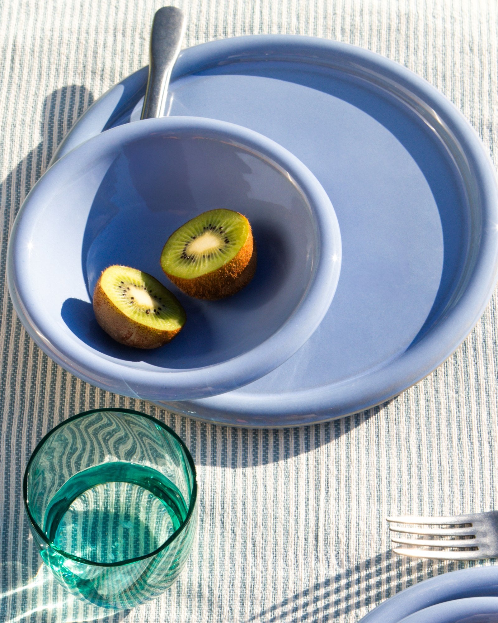 Zwei halbe Kiwis liegen in einer blauen Schüssel auf einem Teller, daneben ein Glas und eine Gabel auf einem gestreiften Tuch.