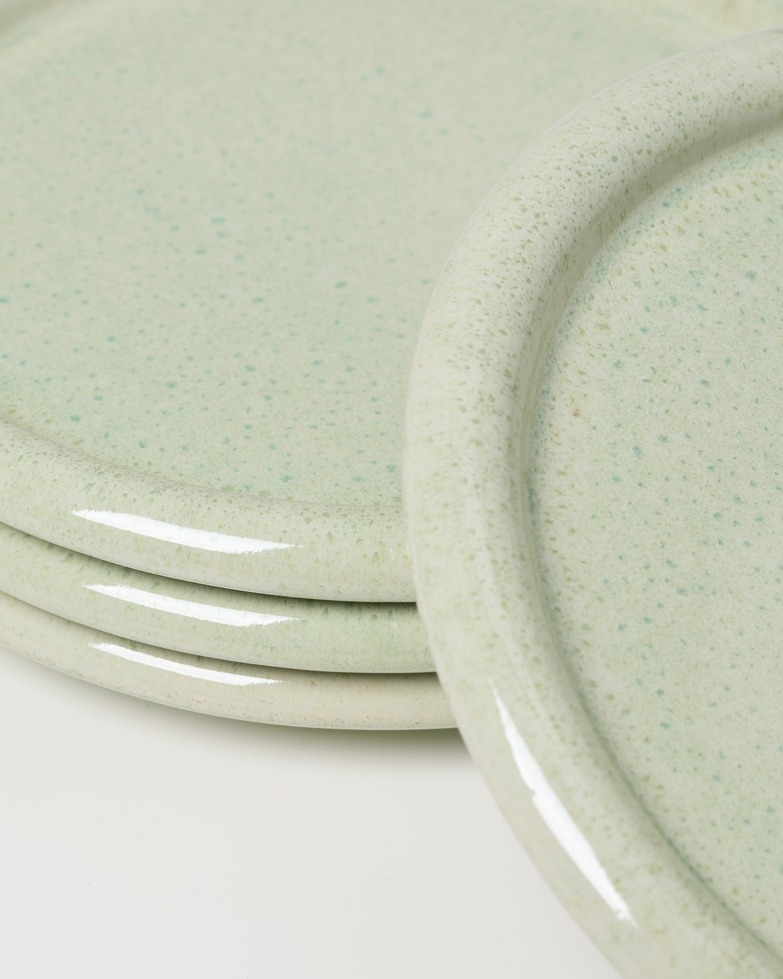 Stapel aus vier hellgrünen, gesprenkelten Keramiktellern mit abgerundeten Kanten auf weißem Hintergrund.