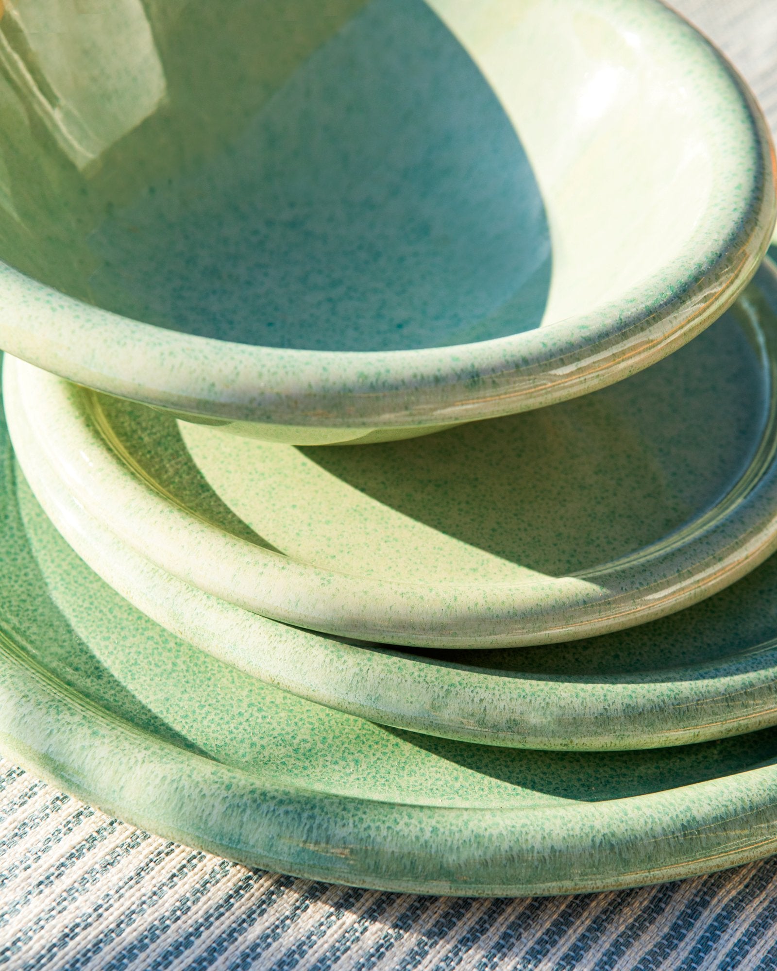 Stapel von grünen, gesprenkelten Keramiktellern auf einem gestreiften Tuch im Sonnenlicht.