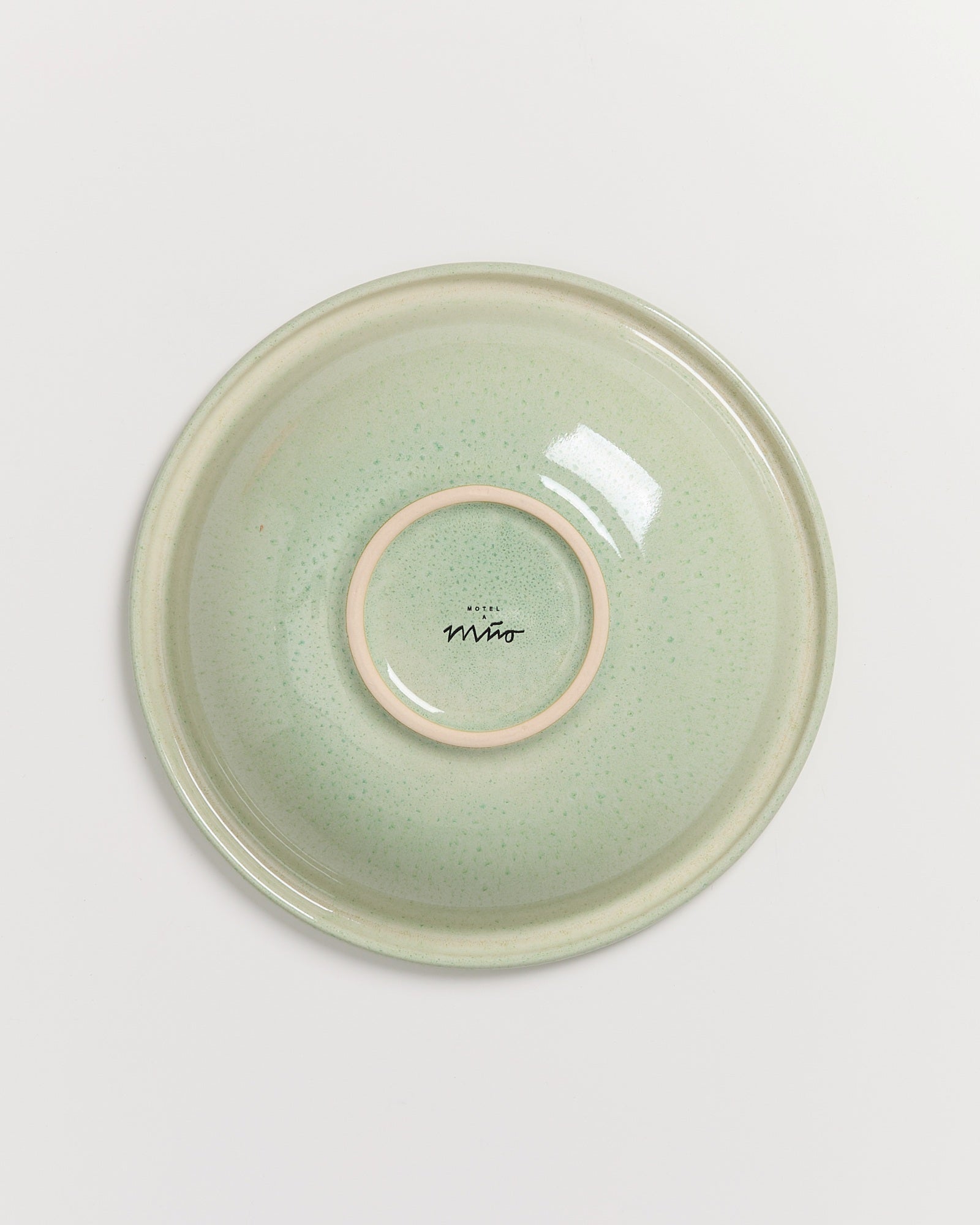 Hellgrüner Keramikteller mit fleckiger Glasur und einem Schriftzug in der Mitte, auf weißem Hintergrund.