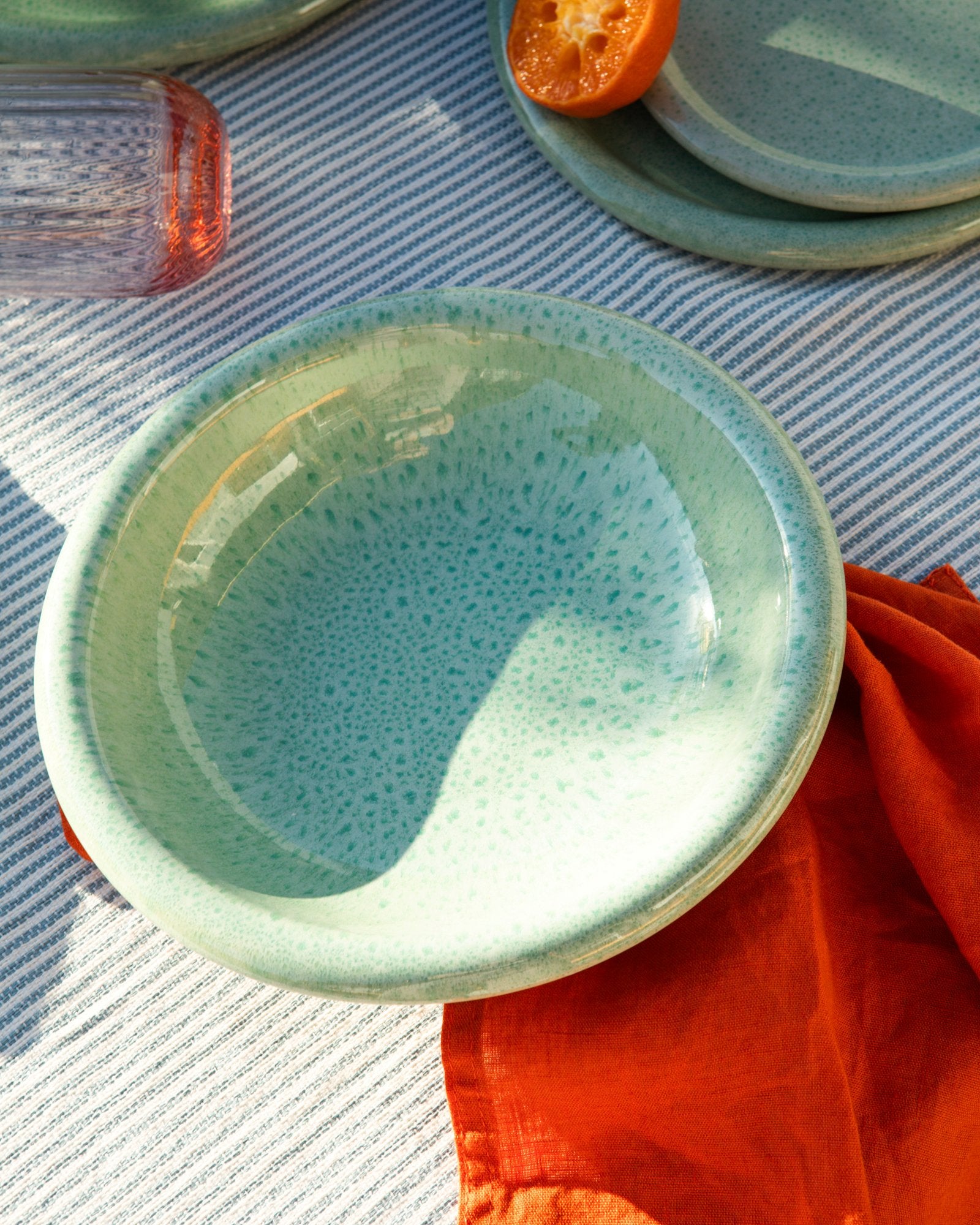 Grüne Keramikschale auf blau-weiß gestreiftem Tuch, mit einer halben Orangenscheibe und einem roten Glas daneben.