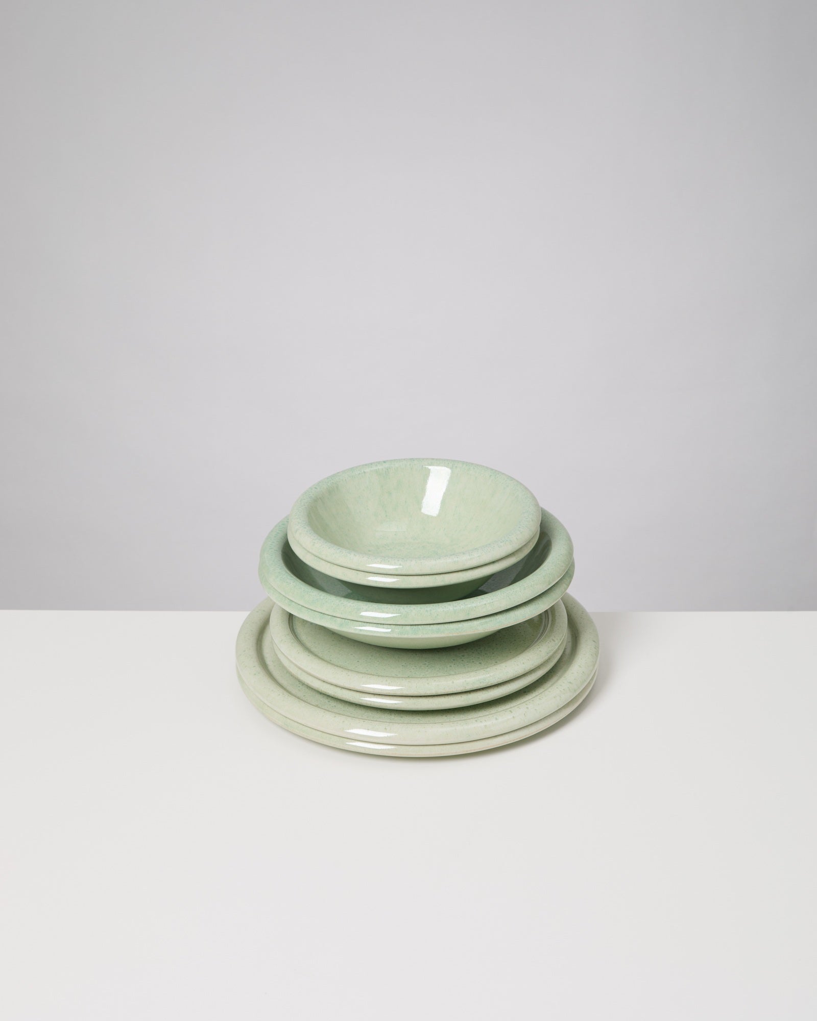 Ein Stapel aus vier grünlichen Keramiktellern und einer Schüssel auf einem weißen Tisch vor grauem Hintergrund.
