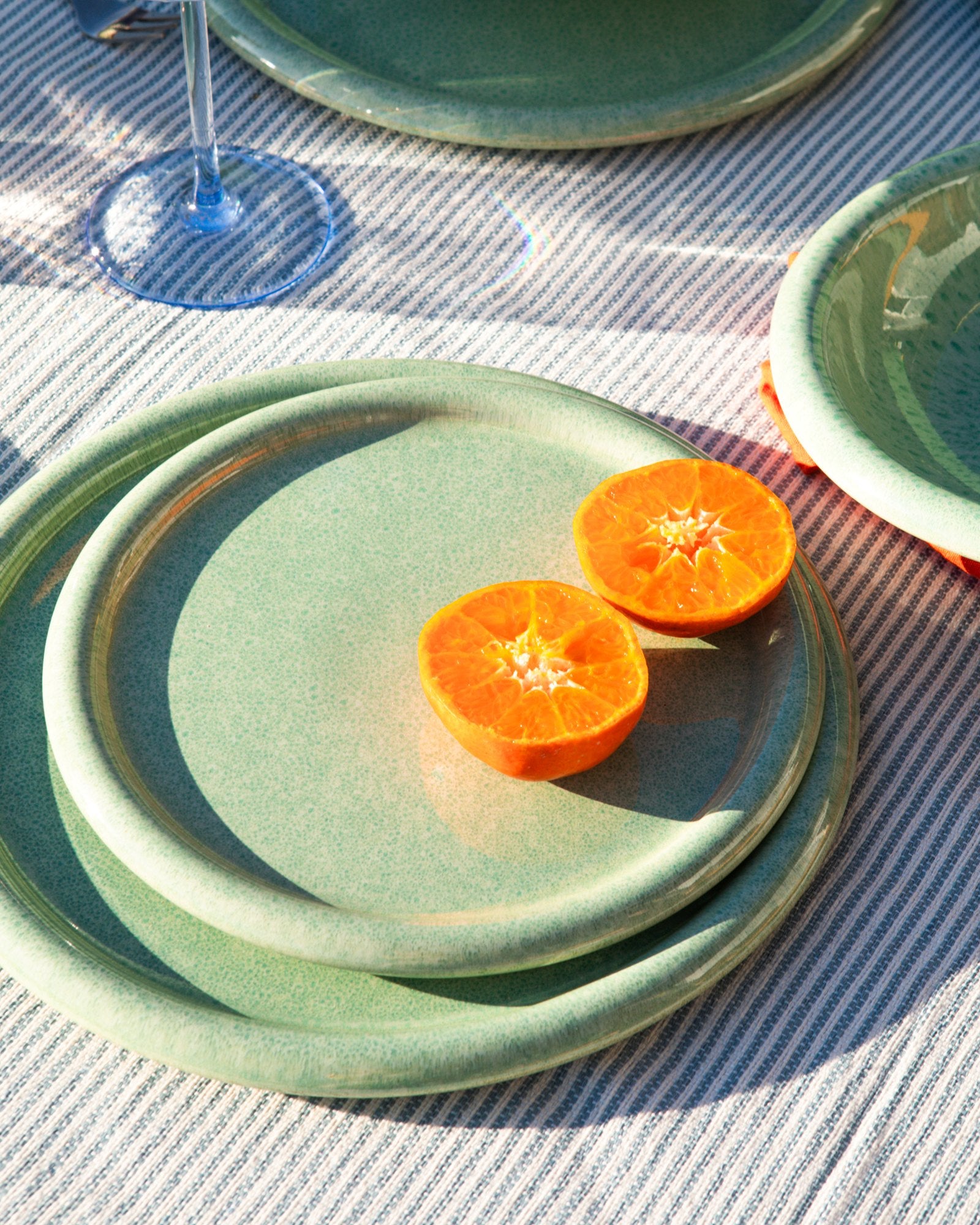 Zwei halbierte Mandarinen auf grünen Tellern, die auf einem gestreiften Tischtuch stehen, mit einem Weinglas daneben.
