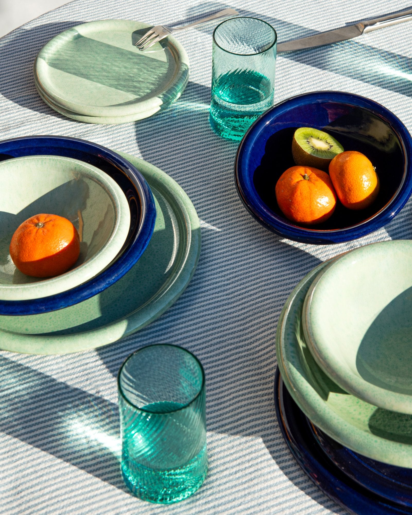 Ein gedeckter Tisch mit grünen und blauen Tellern, Gläsern und Schalen, gefüllt mit Mandarinen und einer Kiwi.