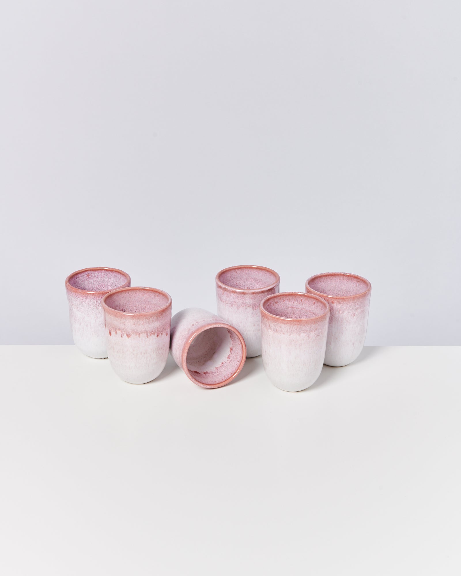 Sechs weiße Keramiktassen mit rosa Rand stehen auf einer weißen Oberfläche, eine liegt umgekippt.
