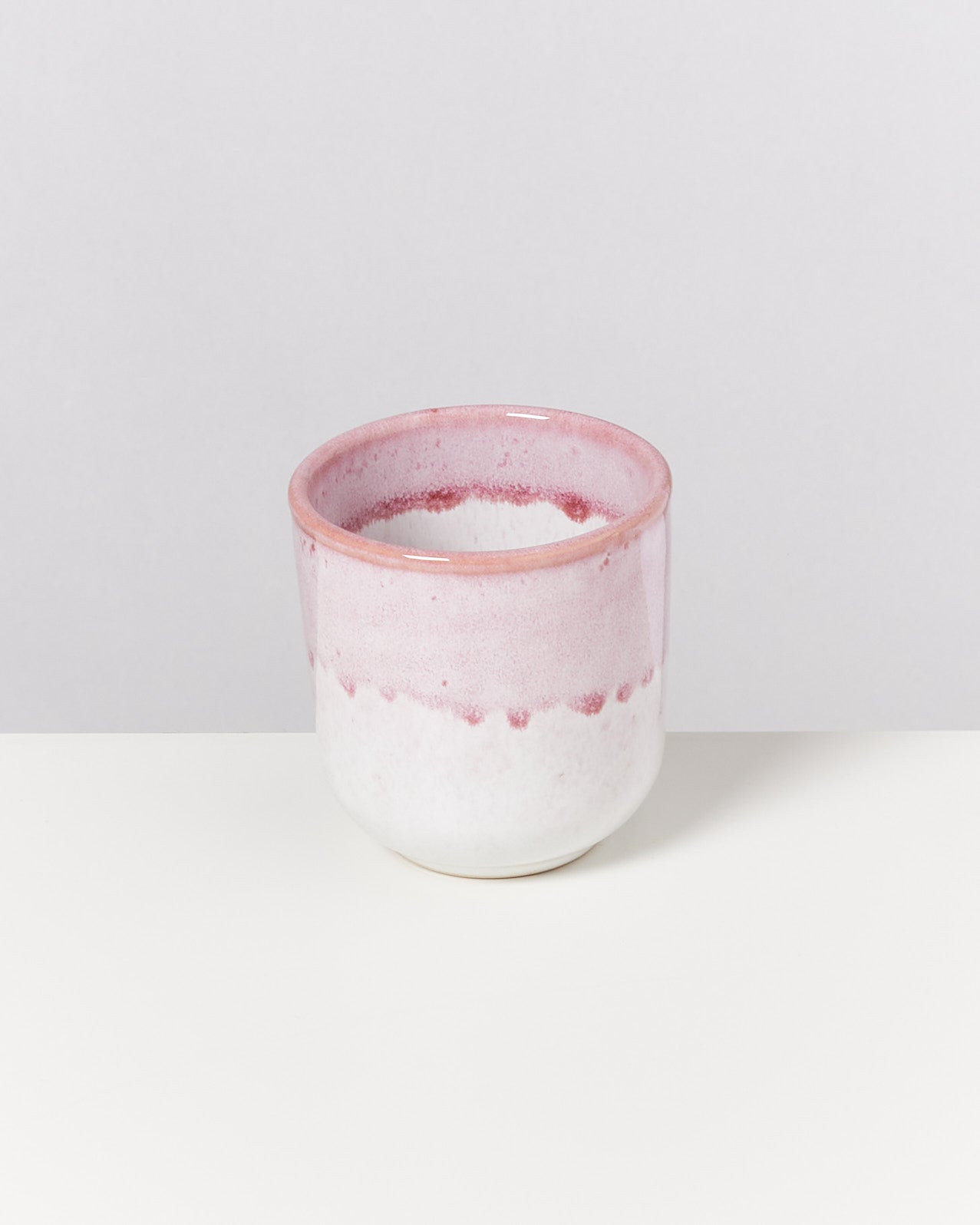 Rosa-weißer Keramikbecher mit glatter Textur, auf einem weißen Untergrund vor einer grauen Wand.