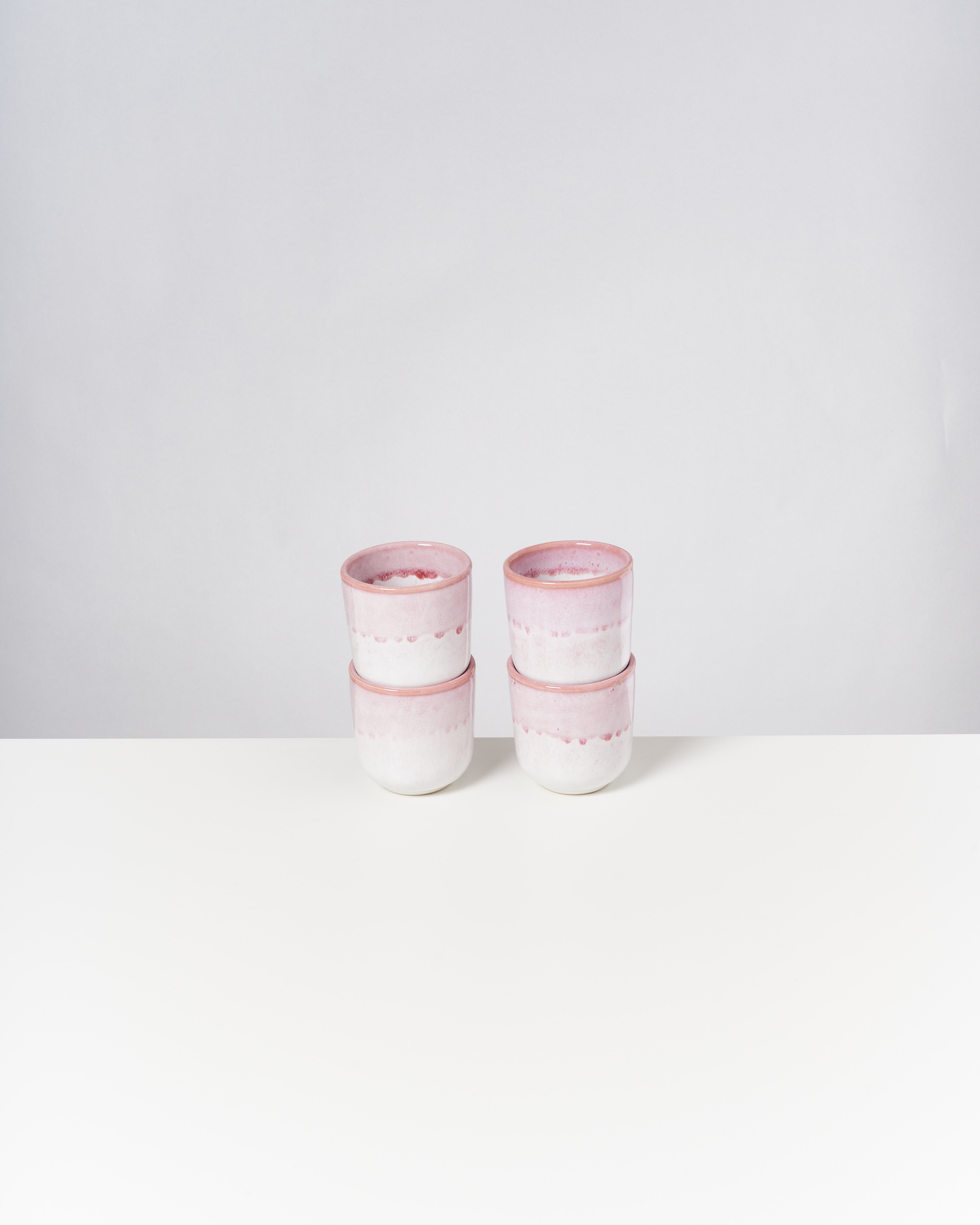 Vier rosafarbene Keramikbecher stehen in zwei Stapeln auf einem weißen Tisch vor einer grauen Wand.