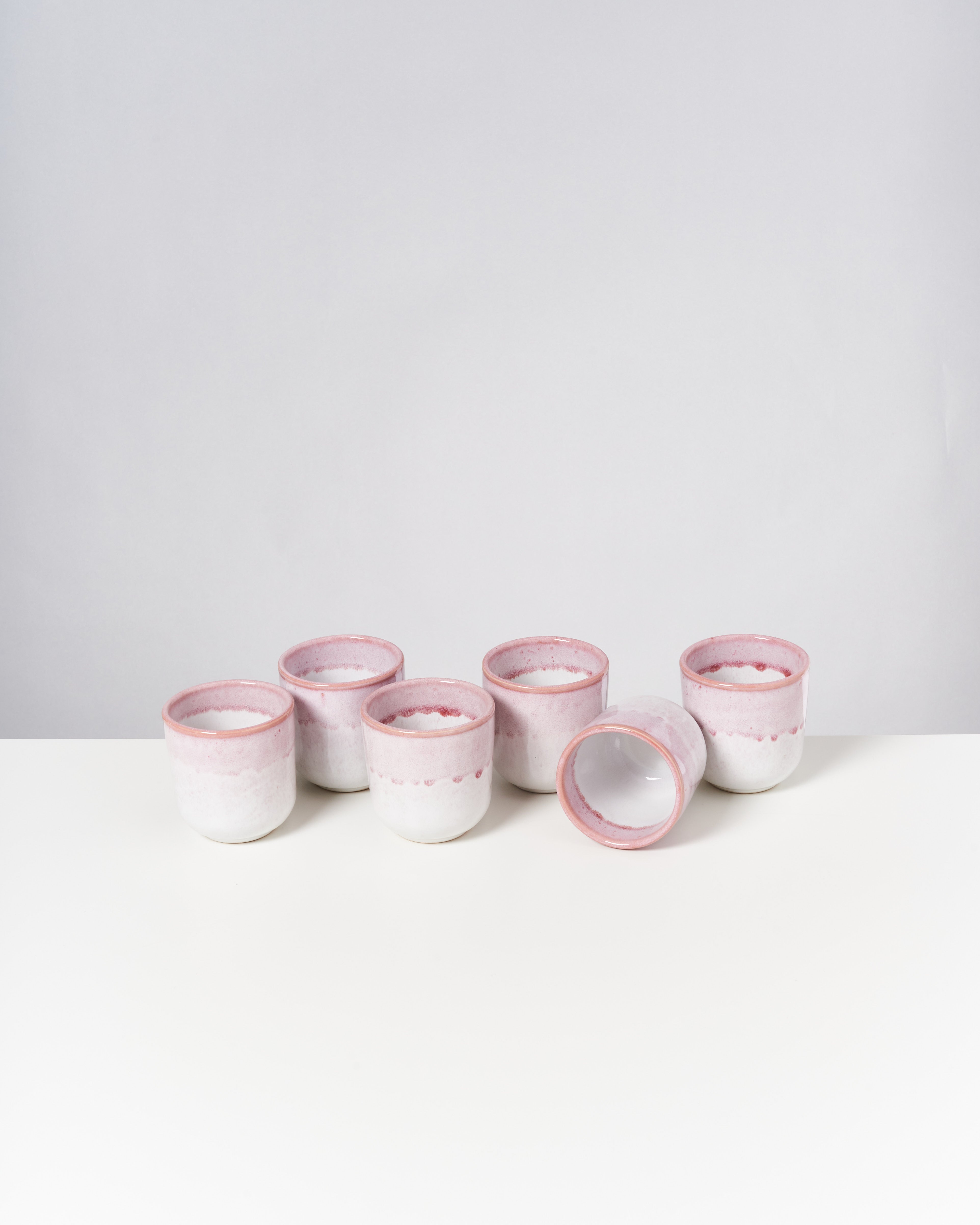Sechs rosa-weiße Keramiktassen, eine liegt auf der Seite, auf einem weißen Tisch vor grauem Hintergrund.