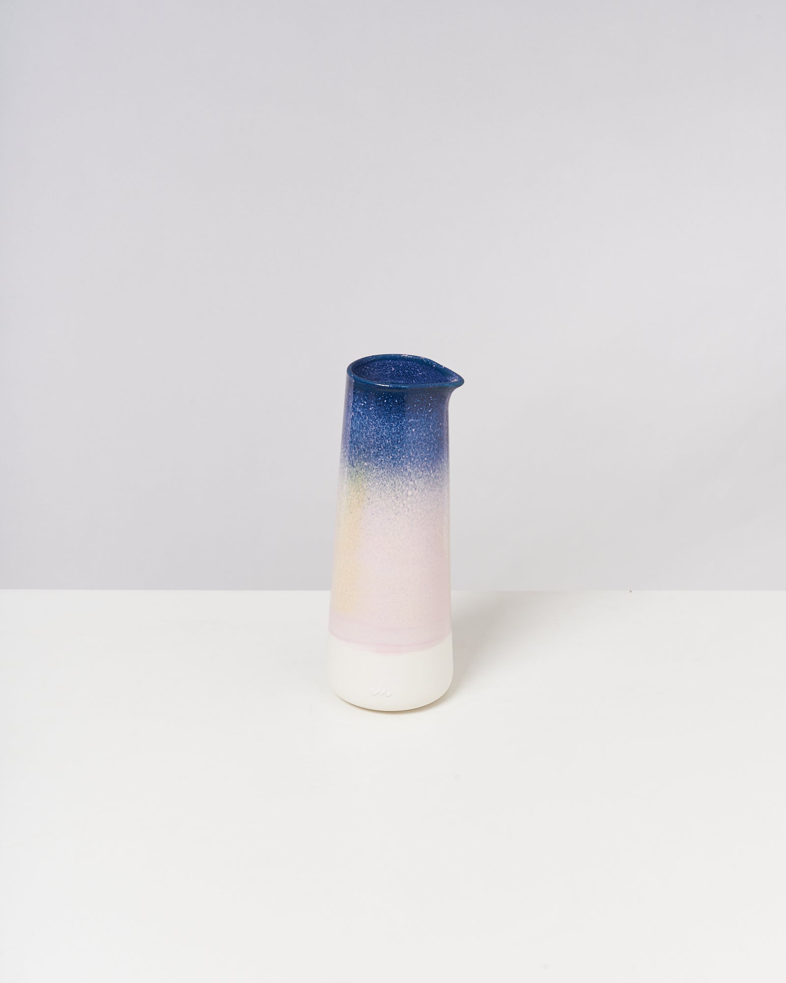 Eine schlanke Vase mit blauem, gelbem und rosafarbenem Farbverlauf steht auf einem weißen Tisch.