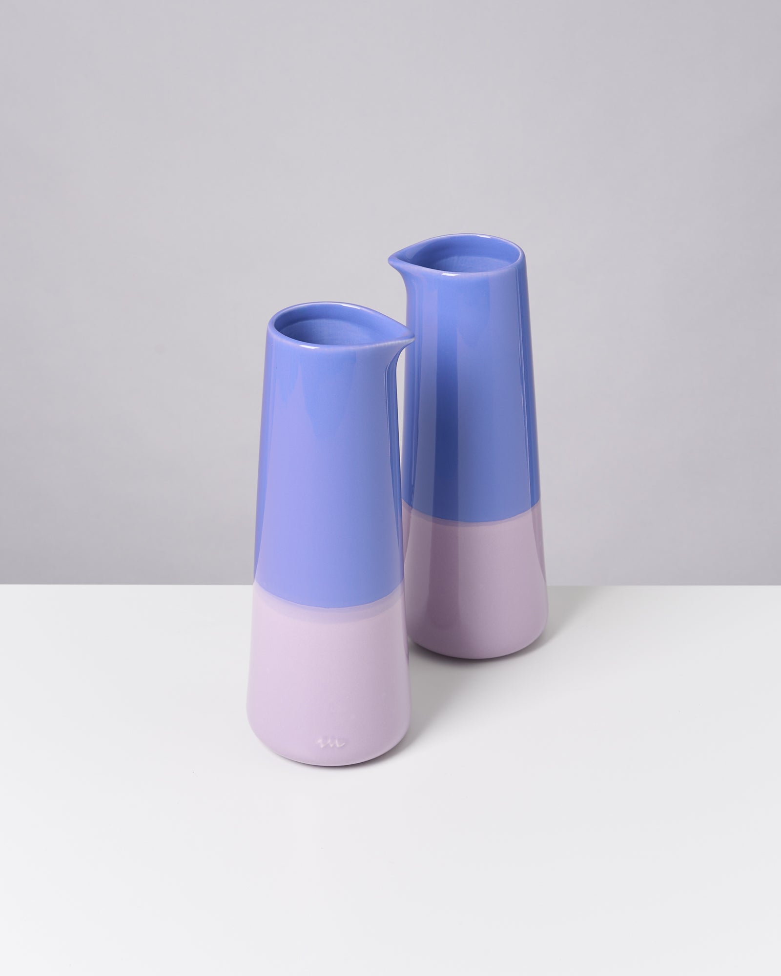 Zwei moderne, zweifarbige Krüge in Lila und Flieder stehen auf einem weißen Tisch vor grauem Hintergrund.