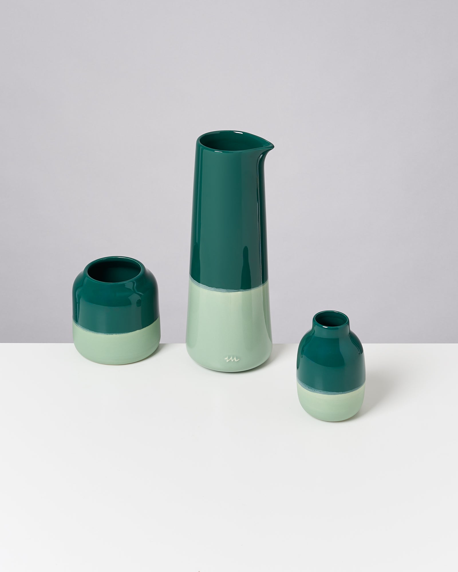 Drei grün-weiße Keramikgefäße in verschiedenen Formen und Größen stehen auf einem weißen Tisch vor grauem Hintergrund.
