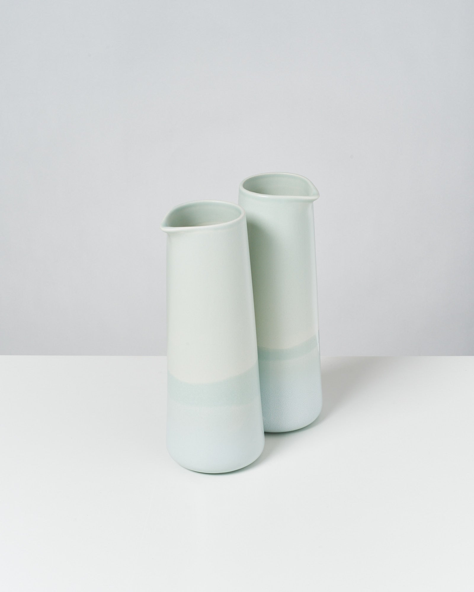 Zwei minimalistische, pastellgrüne Keramikkrüge stehen auf einem weißen Tisch vor einem grauen Hintergrund.