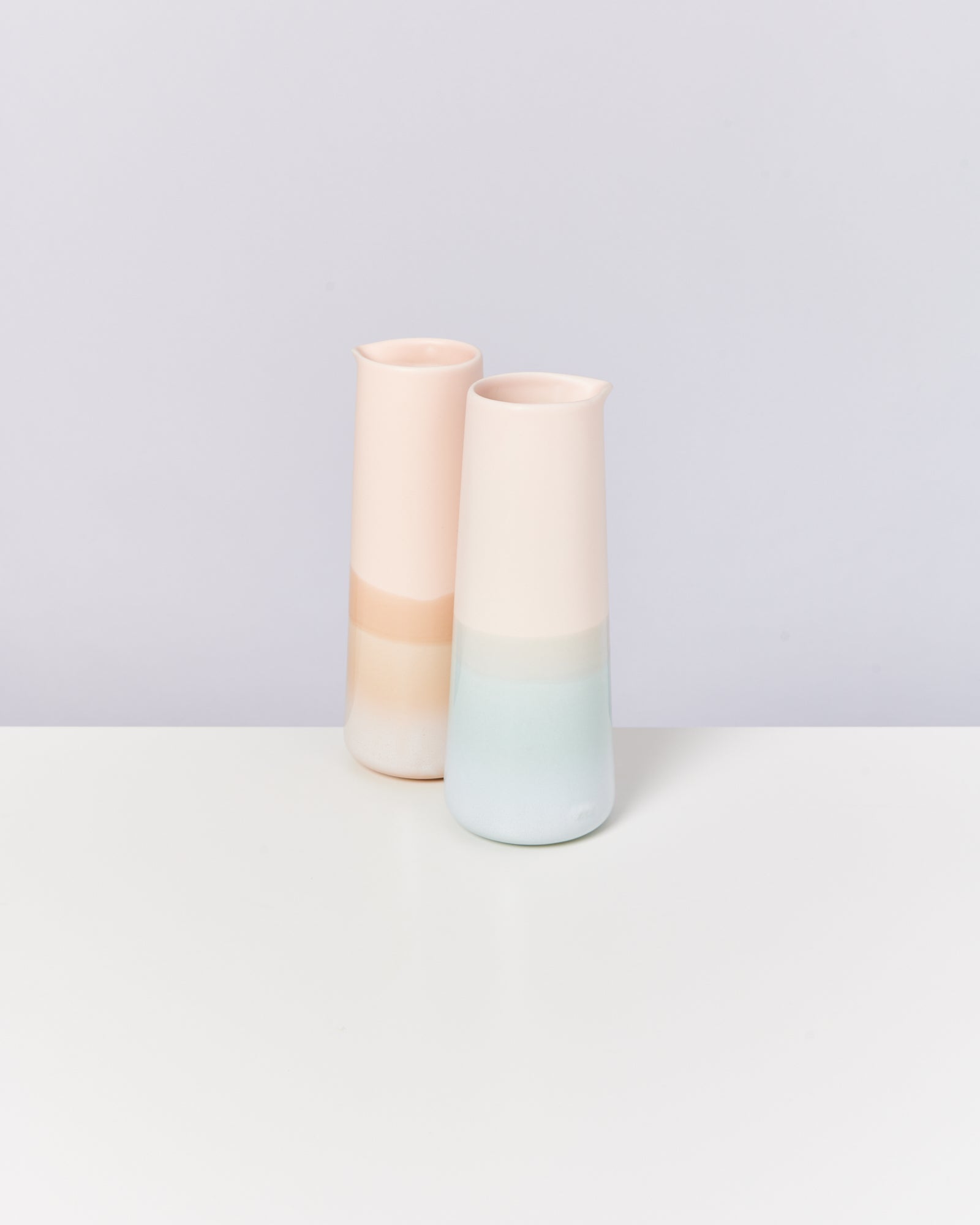 Zwei hohe, pastellfarbene Keramikkrüge stehen auf einem weißen Tisch vor einem grauen Hintergrund.