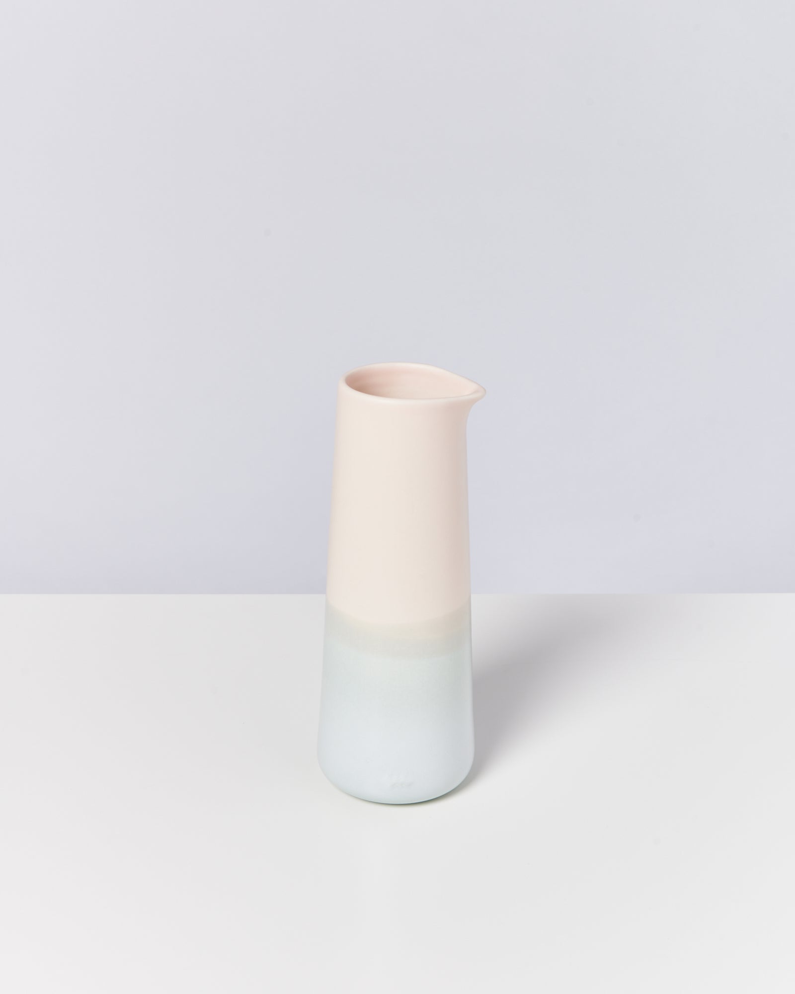 Schlanker, zweifarbiger Krug in Pastellrosa und Blau steht auf einem weißen Tisch vor einem grauen Hintergrund.