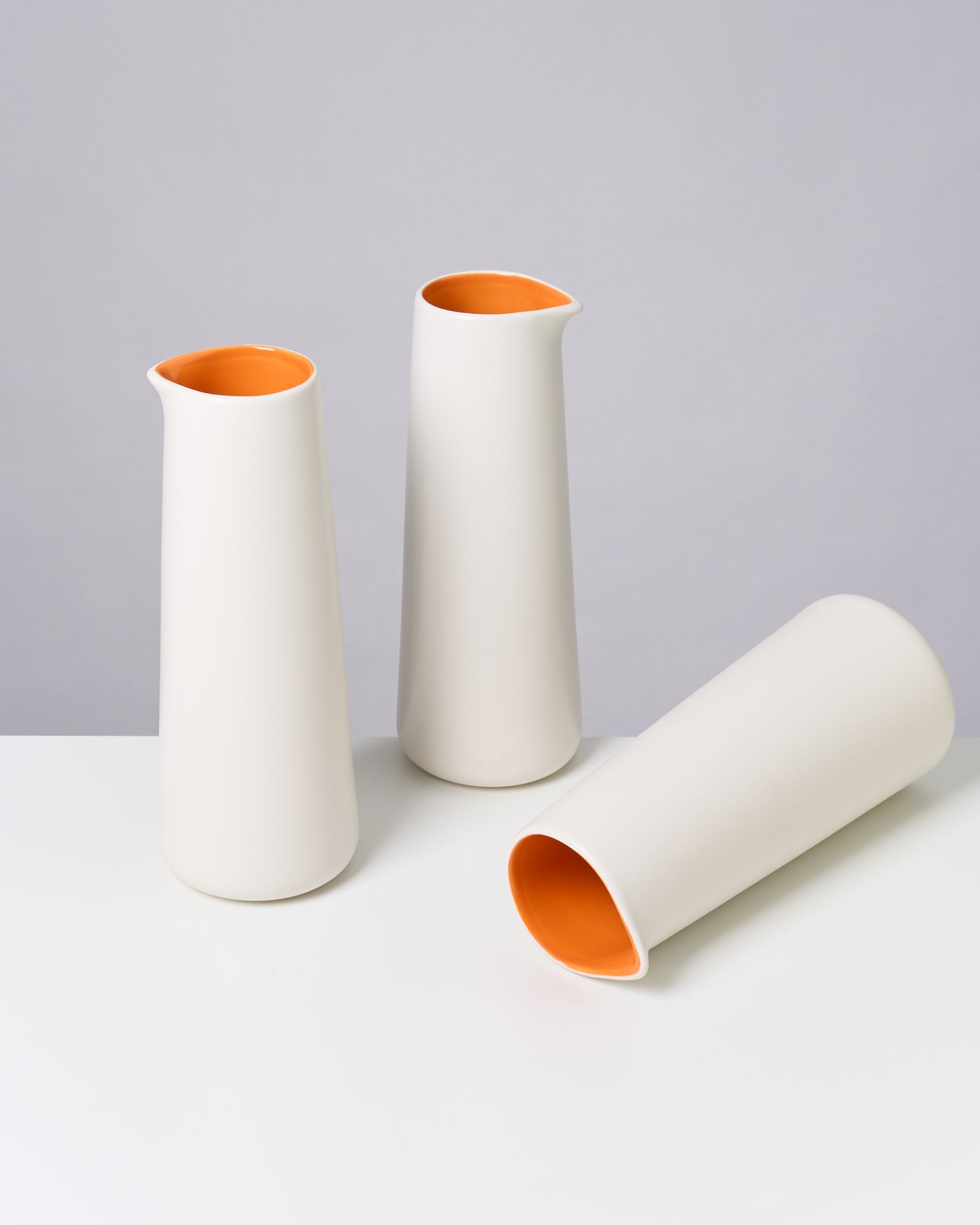 Drei weiße Vasen mit orangefarbenem Inneren stehen auf einem weißen Tisch, eine liegt auf der Seite.