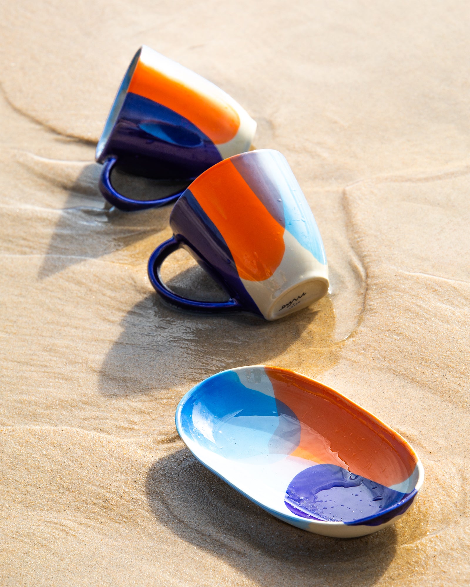 Zwei bunte Tassen und ein passender Teller liegen auf nassem Sand am Strand.