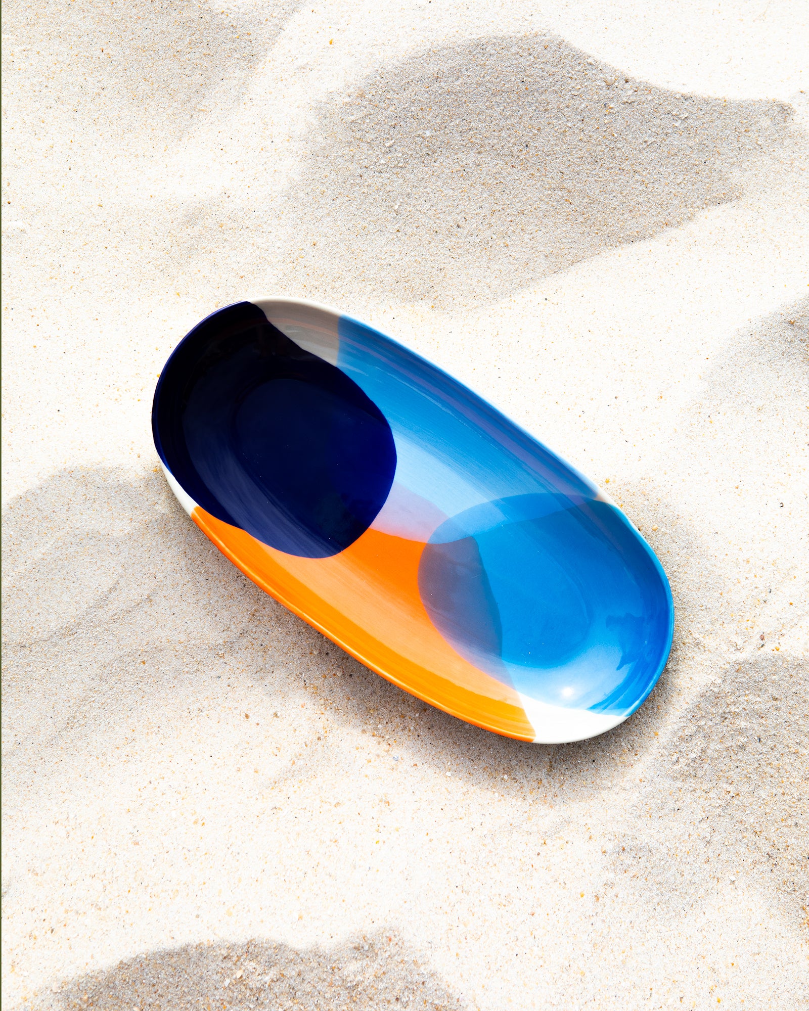 Bunte ovale Schale mit Blau-, Weiß- und Orangetönen liegt auf hellem Sand.