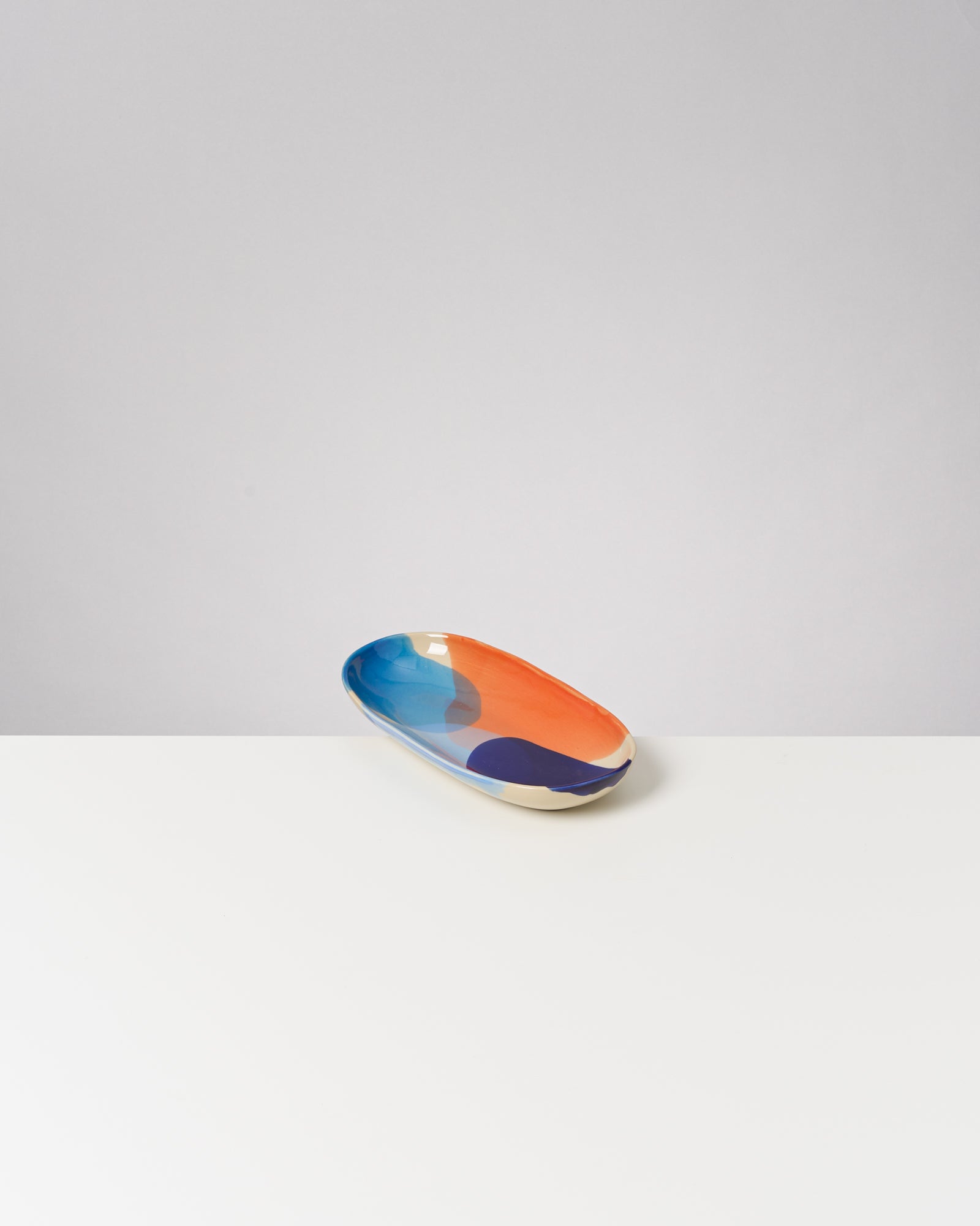 Langes, ovales Keramiktablett mit blau-orangefarbenem Muster auf einem weißen Tisch vor grauem Hintergrund.