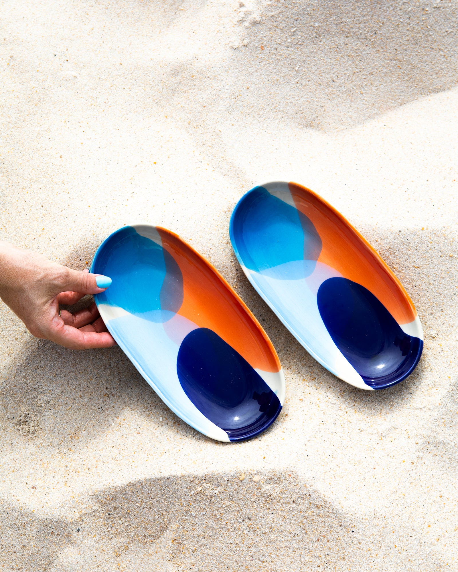 Zwei bunt bemalte Teller liegen auf Sand, während eine Hand einen Teller anhebt.