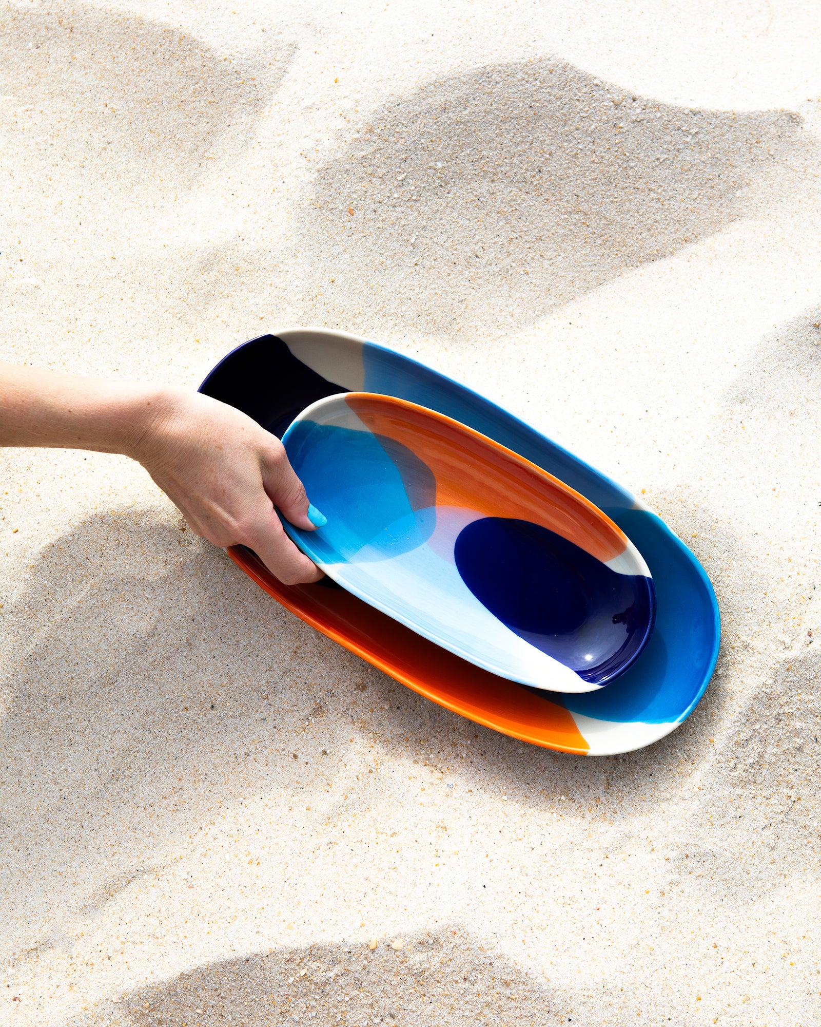 Eine Hand hält zwei bunt bemalte, ovale Teller auf sandigem Untergrund.