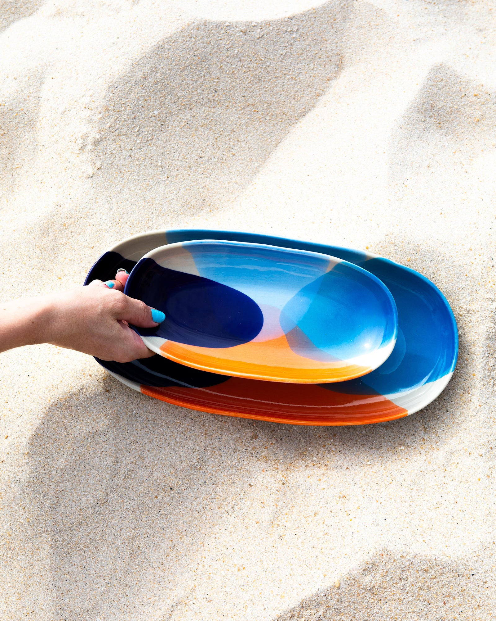 Eine Hand mit blau lackierten Nägeln hält einen bunten Teller auf sandigem Untergrund.