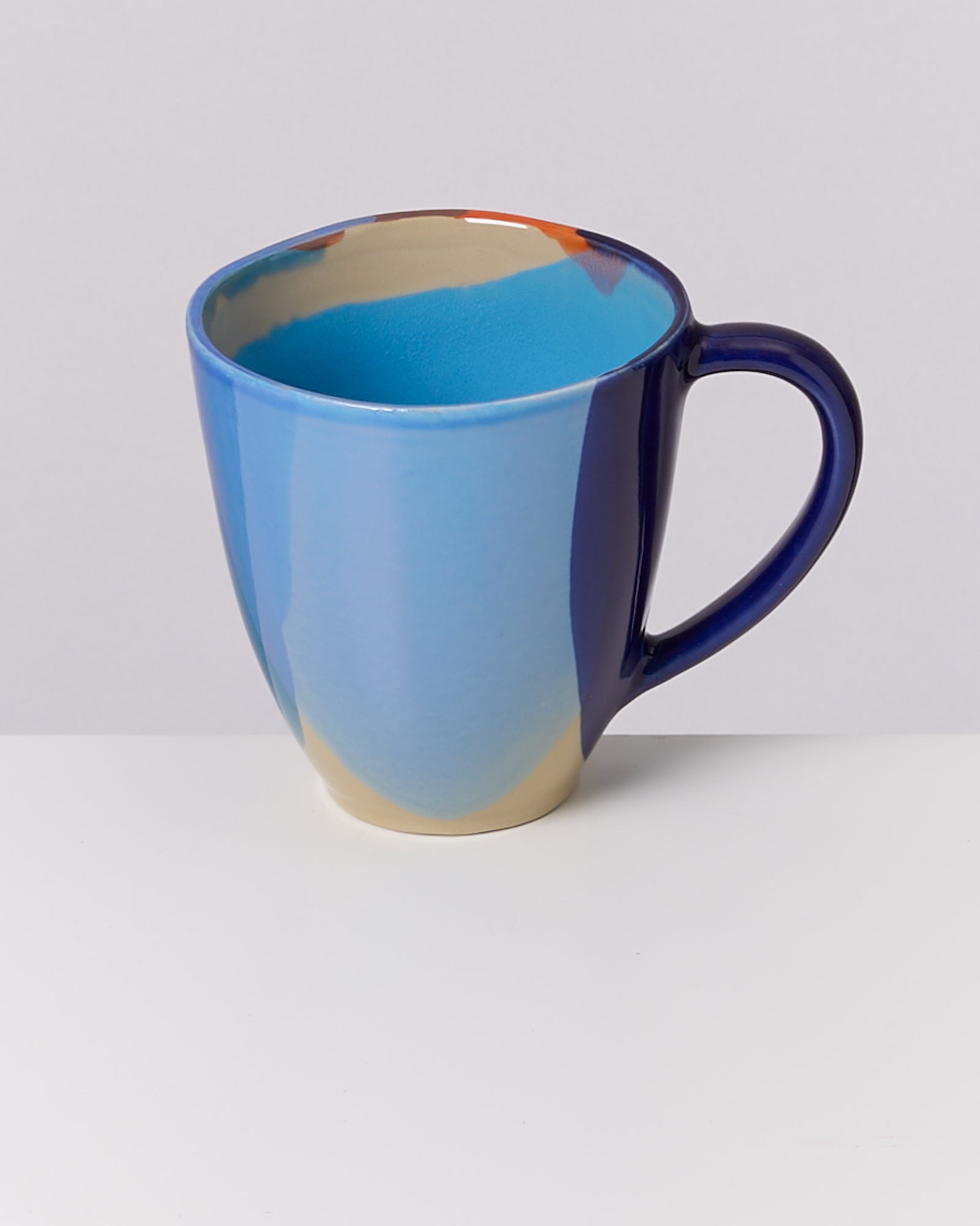 Eine blau-ockerfarbene Keramiktasse mit Henkel auf einem weißen Untergrund und grauem Hintergrund.