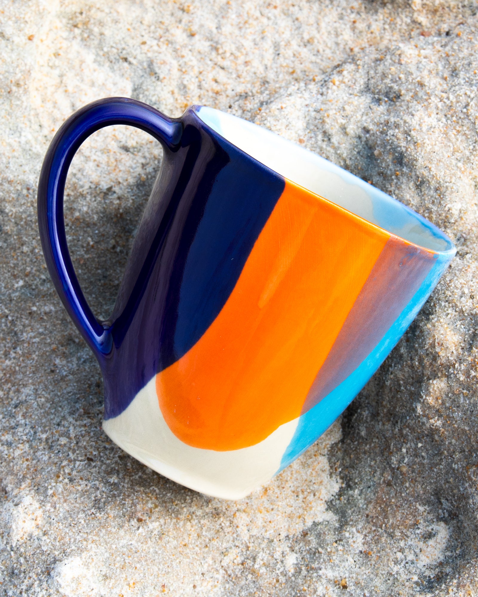 Bunte Kaffeetasse mit blauen, orangen und weißen Streifen liegt auf einem sandigen Untergrund.