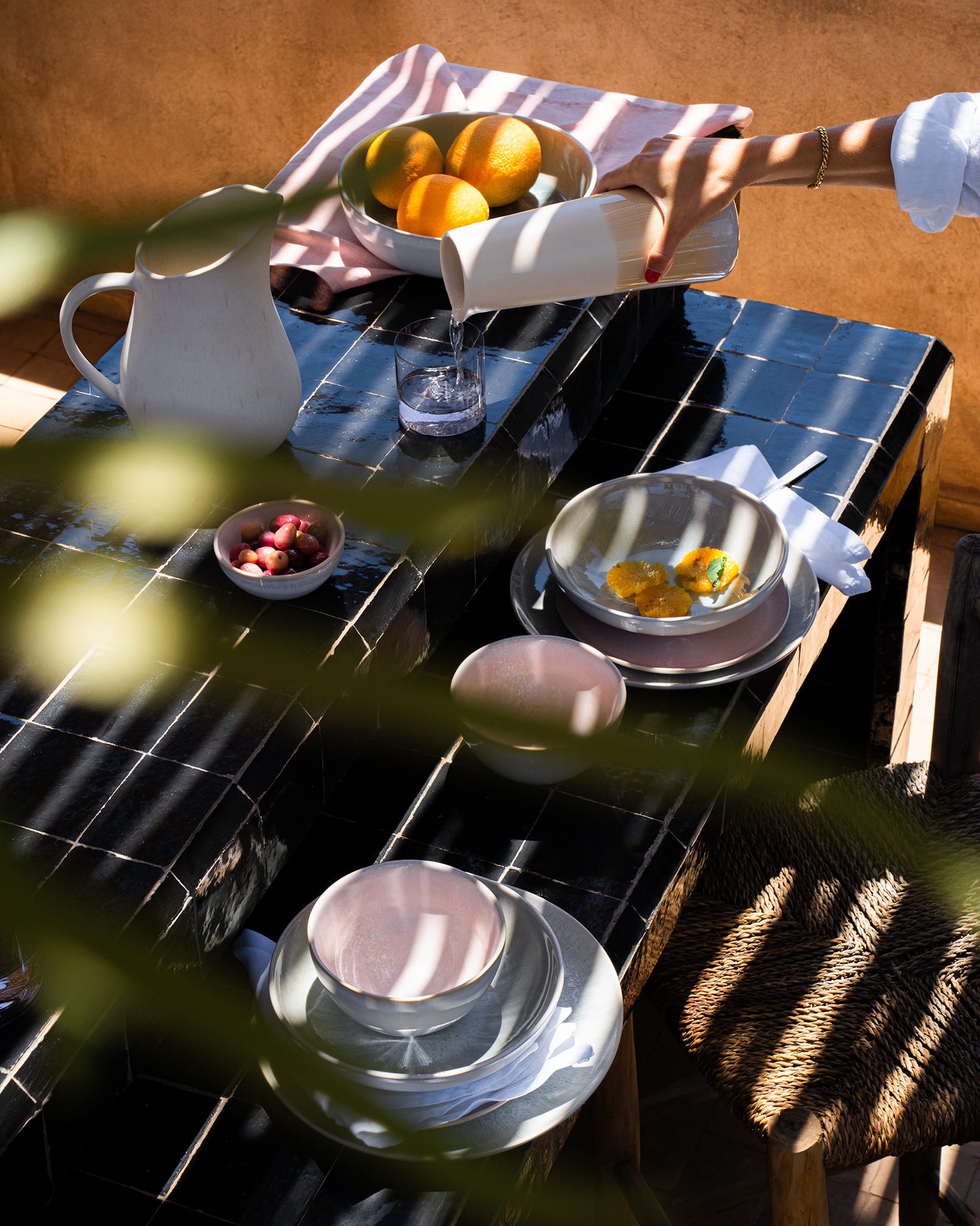 Ein Tisch mit Schalen, einer Kanne, Geschirr und Früchten im Sonnenlicht, jemand gießt Wasser in ein Glas.