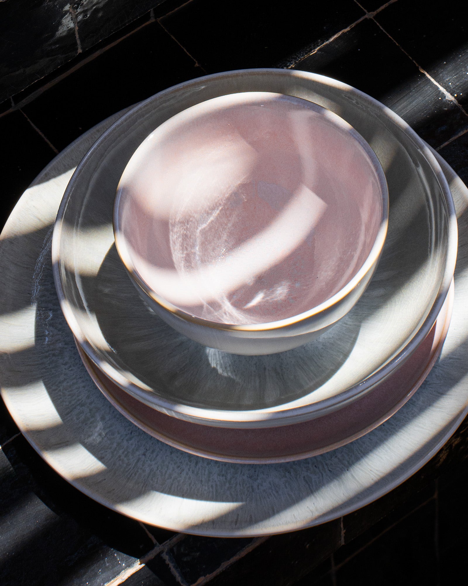 Stapel von Tellern und einer Schüssel in sanften Pastellfarben, beleuchtet von Sonnenlicht, auf einer dunklen Oberfläche.