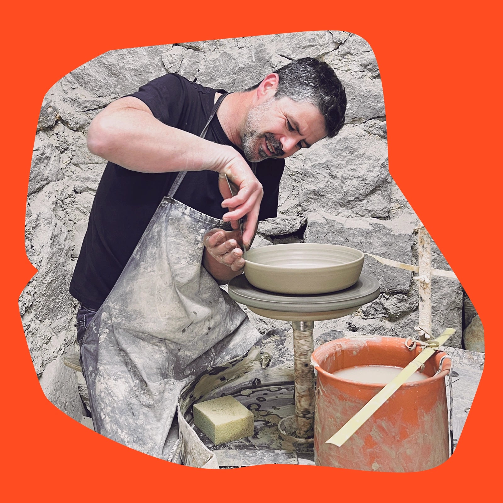 Ein Mann arbeitet an einer Töpferscheibe, umgeben von Werkzeugen und Materialien, vor einem Stein-Hintergrund.