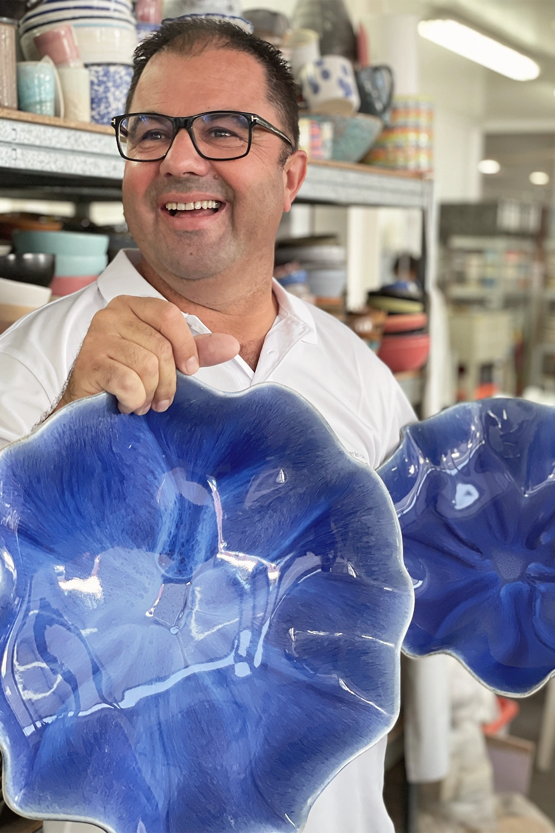 Mann mit Brille im weißen Hemd hält zwei große, blaue Keramikteller und lächelt in einem Laden mit vielen Geschirrartikeln.
