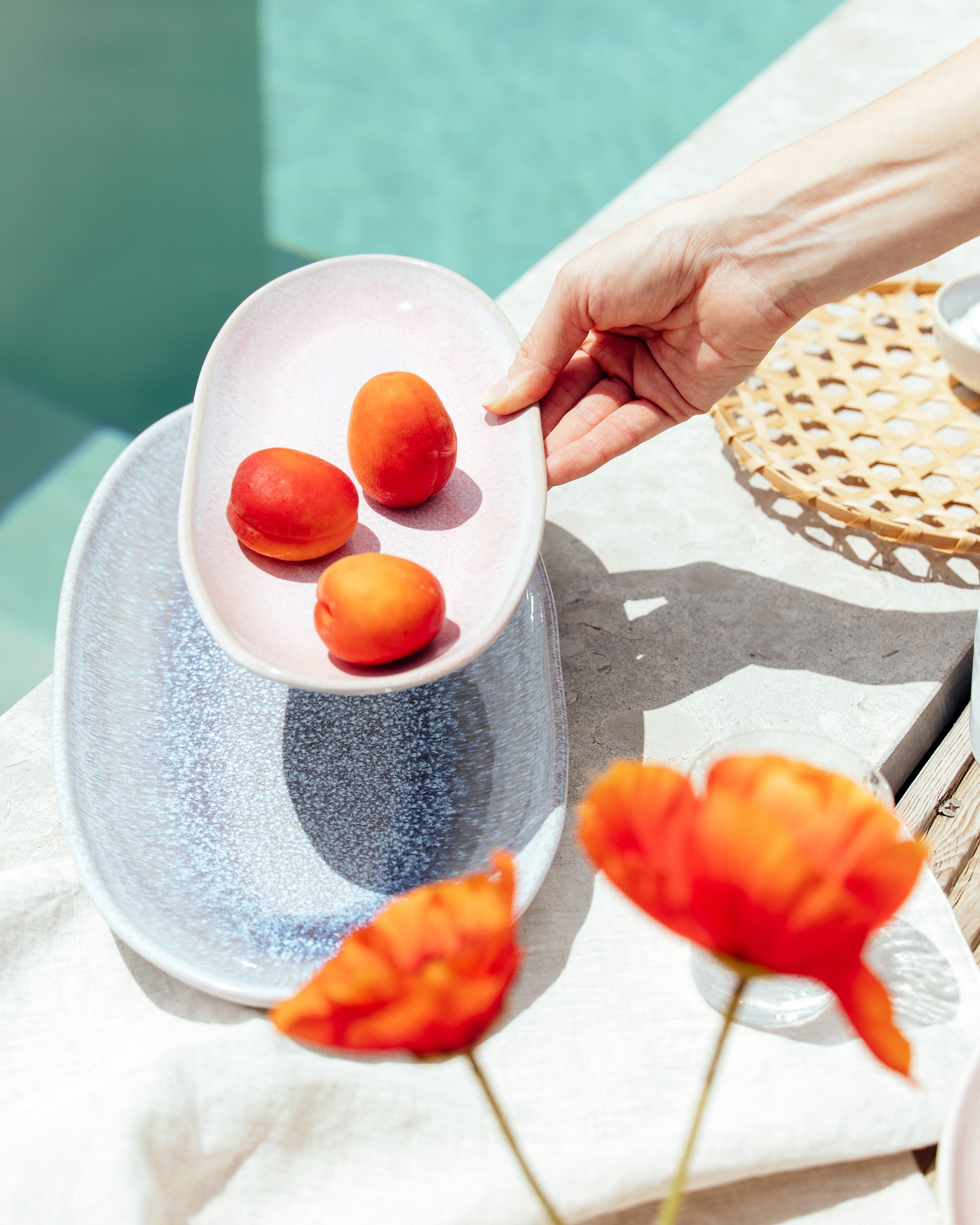 Eine Hand hält einen Teller mit Aprikosen über einem größeren Teller am Rand eines Pools, im Vordergrund sind orangefarbene Blumen.