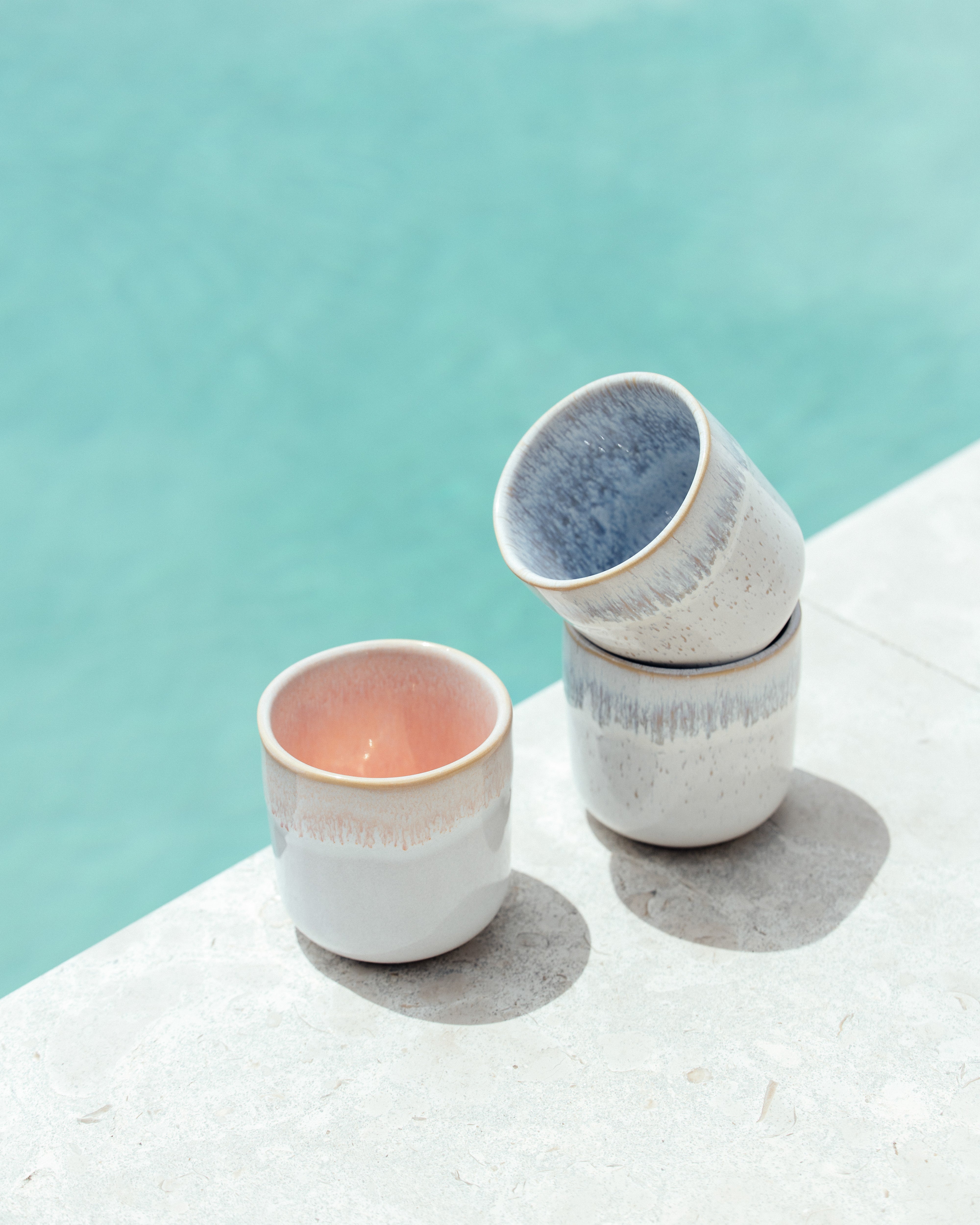 Zwei gestapelte Keramikbecher und ein einziger Becher auf einem hellen Steinboden, mit einem Pool im Hintergrund.