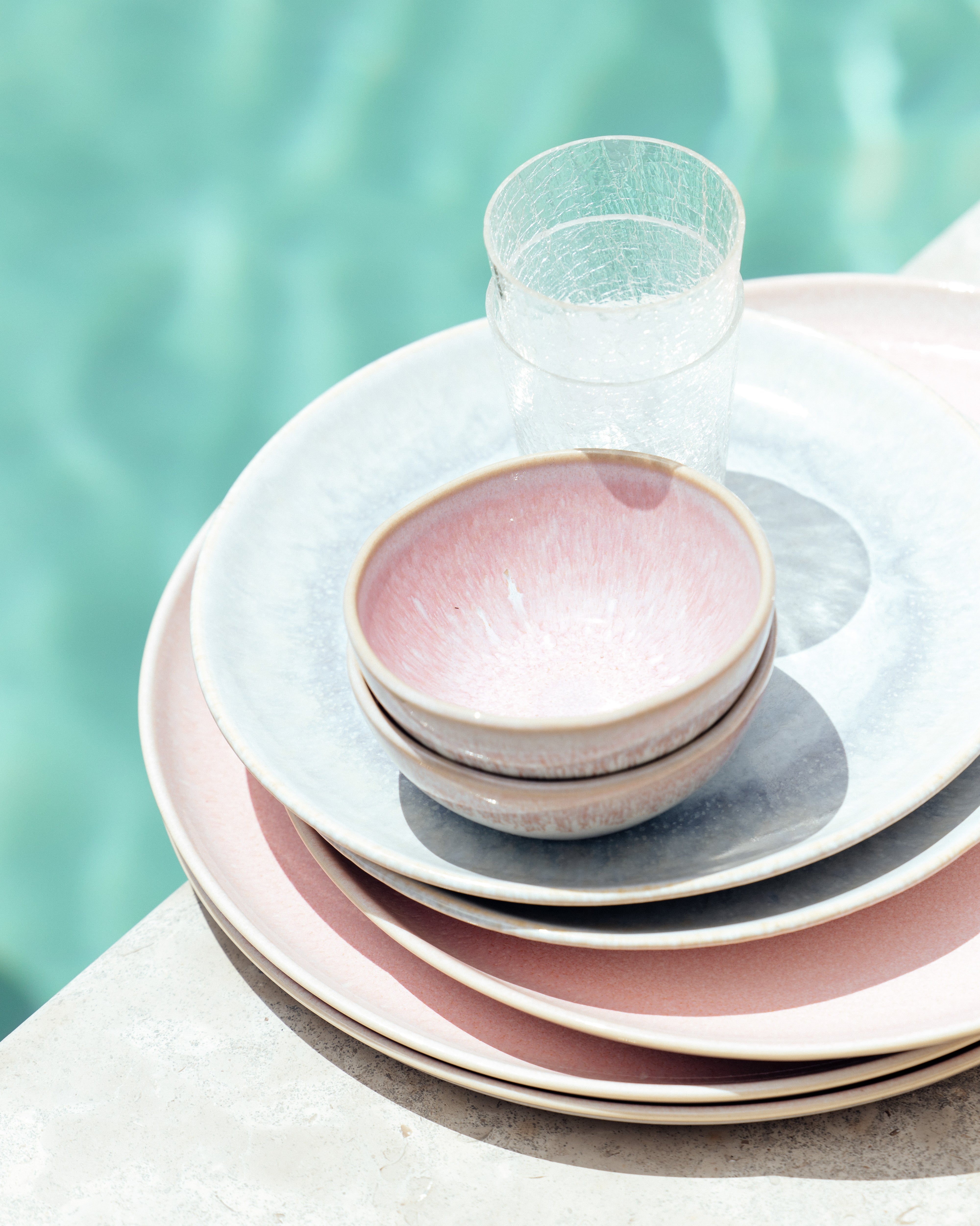 Gestapelte Teller und Schalen in Pastellfarben neben einem klaren Glas auf marmorierter Oberfläche, mit Pool im Hintergrund.