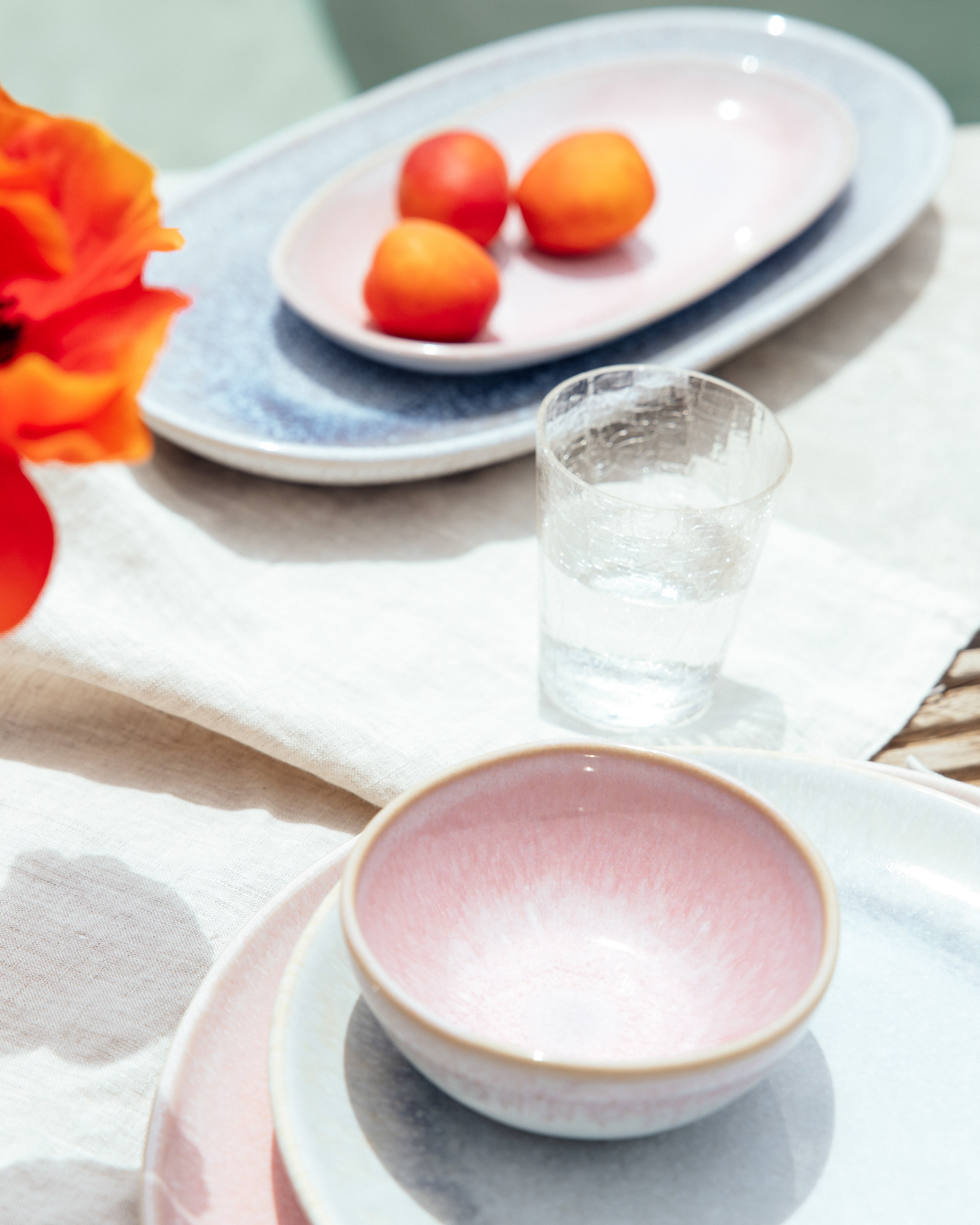 Tisch mit rosa und blauen Keramikschalen, einem Glas Wasser, oranger Blume und Aprikosen auf einem Tablett.