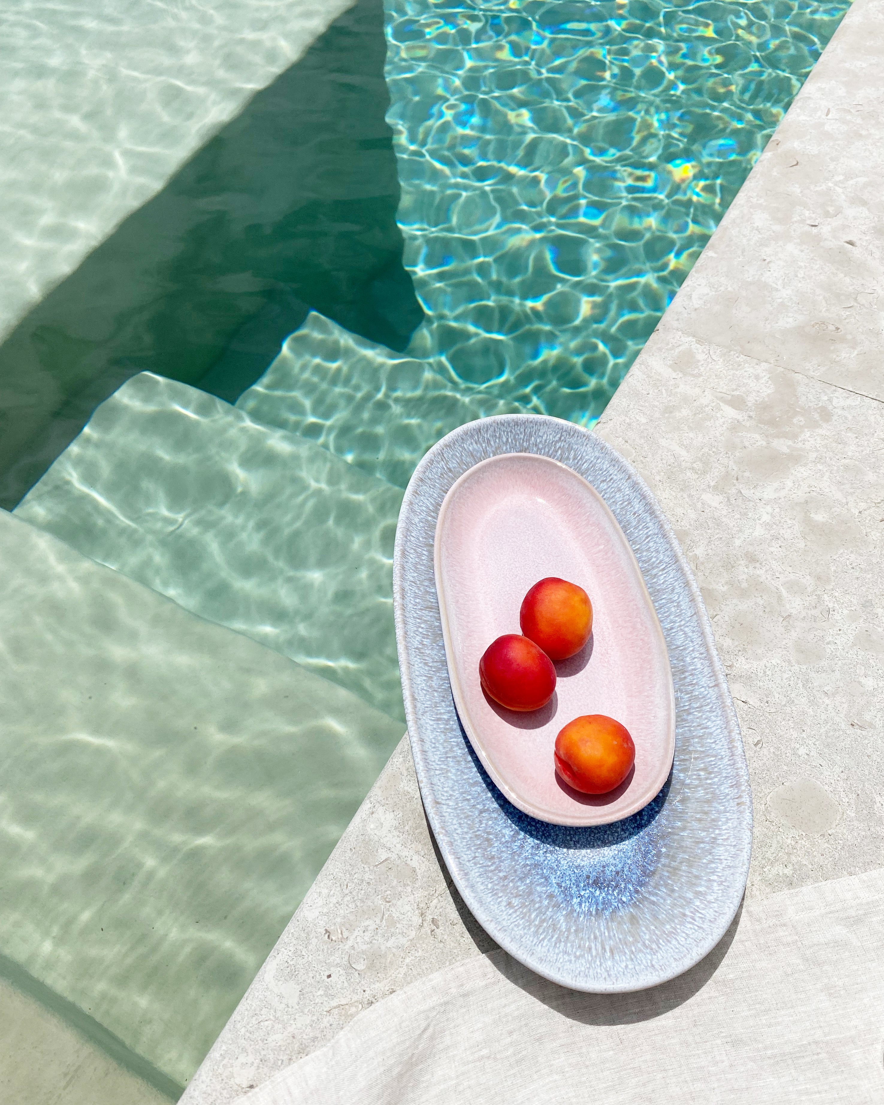 Drei Pfirsiche liegen auf einem Teller neben einem klaren Swimmingpool auf hellem Steinboden.