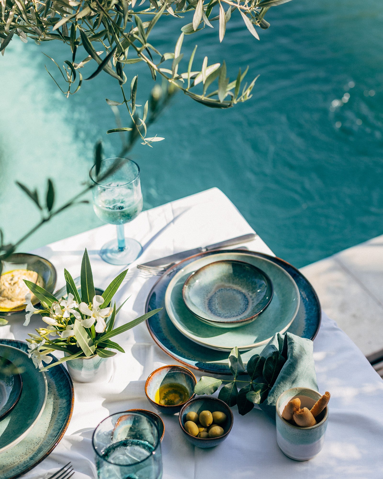 Ein elegant gedeckter Tisch mit blaugrünem Geschirr, Blumen und Oliven nahe eines Pools, umgeben von Olivenzweigen.