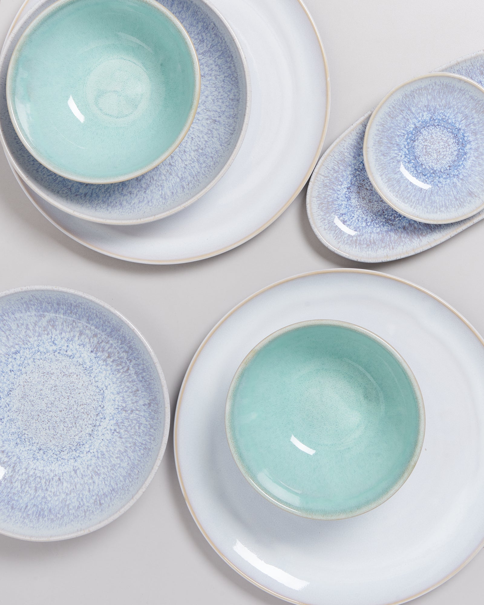 Verschiedene Keramikteller und Schalen in Blau- und Grüntönen liegen auf einer hellen Fläche arrangiert.
