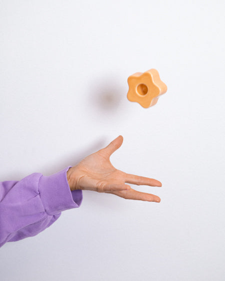 Eine Hand in einem lila Pullover wirft ein orangefarbenes, sternförmiges Objekt in die Luft vor einem weißen Hintergrund.