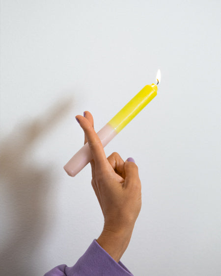 Eine Hand hält eine brennende gelb-rosa Kerze vor einem weißen Hintergrund.