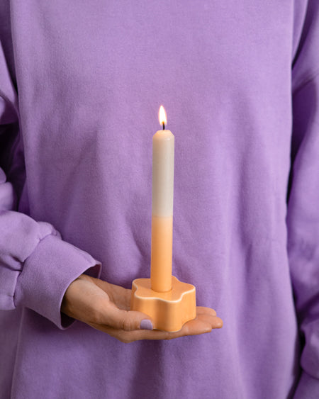 Eine Person in lila Pullover hält eine brennende Kerze in einem orangefarbenen Kerzenhalter.