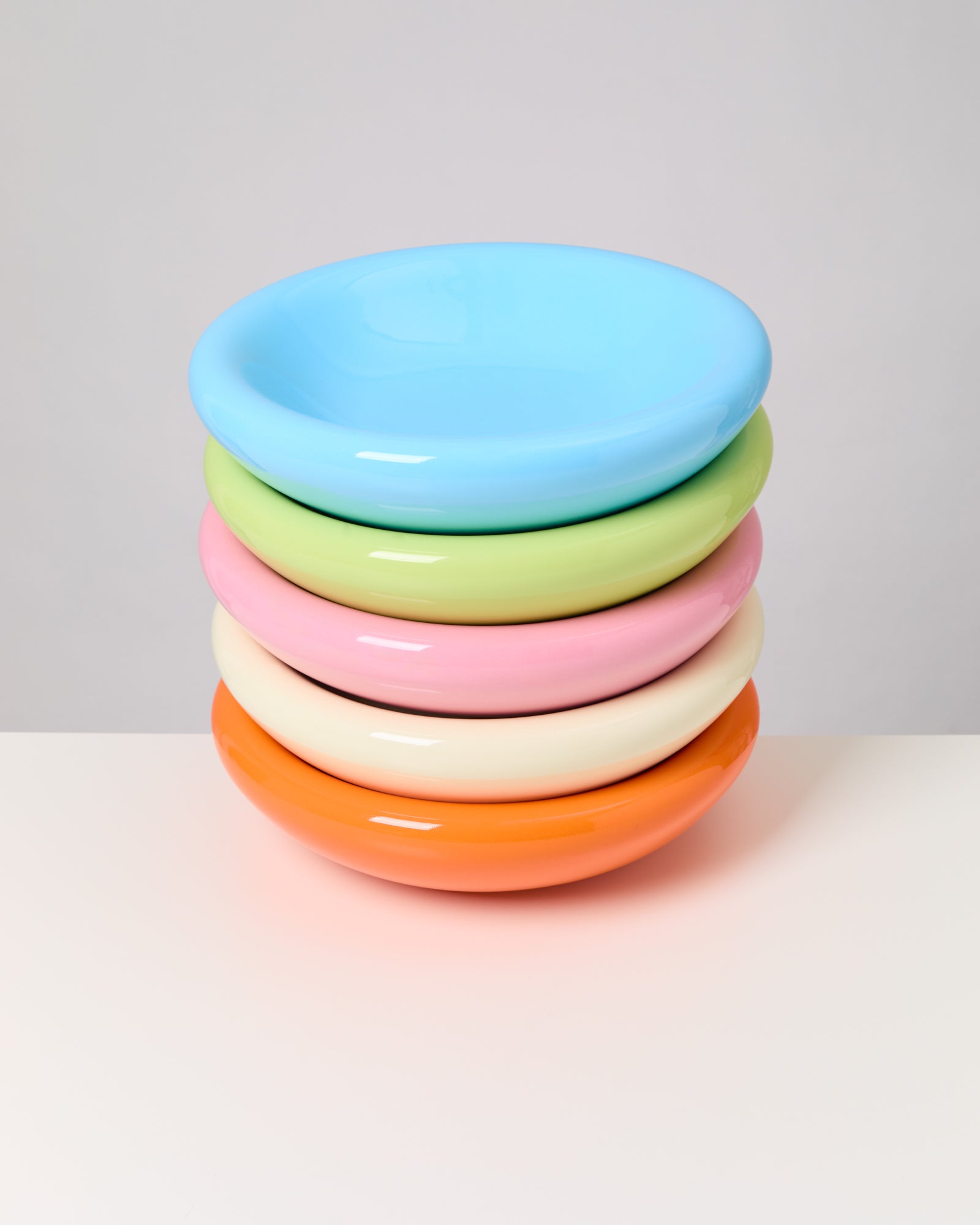 Fünf bunte, pastellfarbene Schalen sind übereinandergestapelt auf einem weißen Tisch vor grauem Hintergrund.