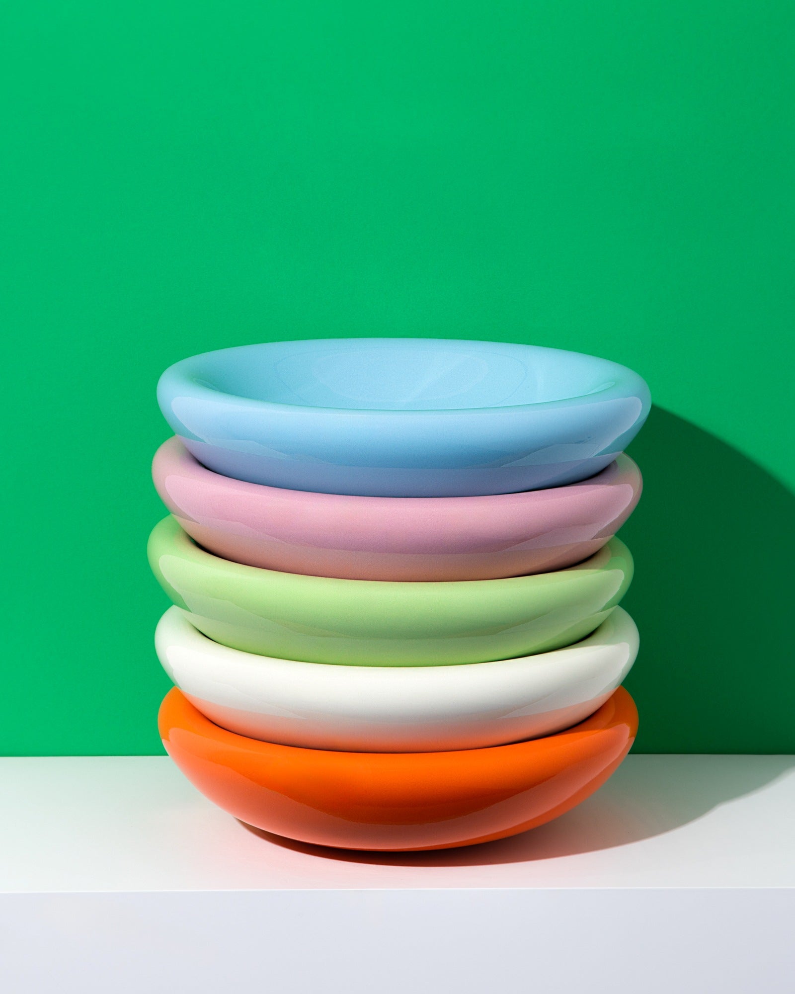 Ein Stapel von sechs bunten Tellern auf einem weißen Tisch vor grüner Wand.