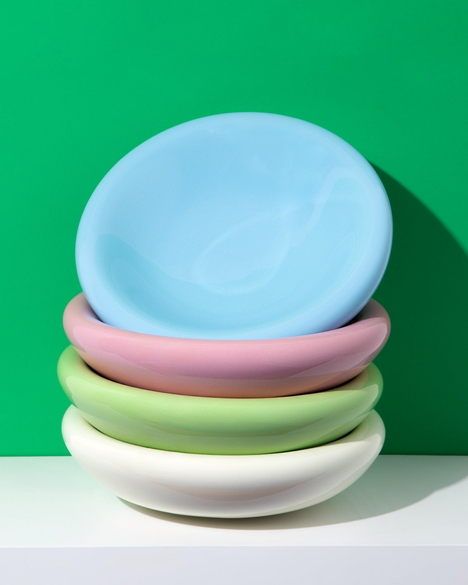 Ein Stapel bunter, pastellfarbener Teller vor grünem Hintergrund auf einem weißen Regal.